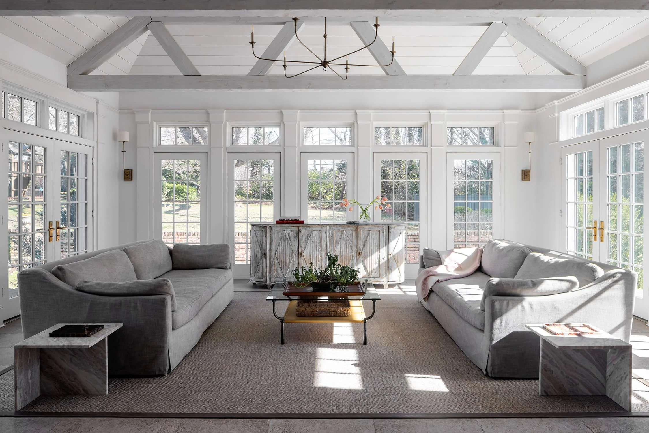 Sunroom with white painted exposed truss ceiling, twin sofas, and wall of French doors overlooking the garden, photographed by Brian Berkowitz Photography on Long Island