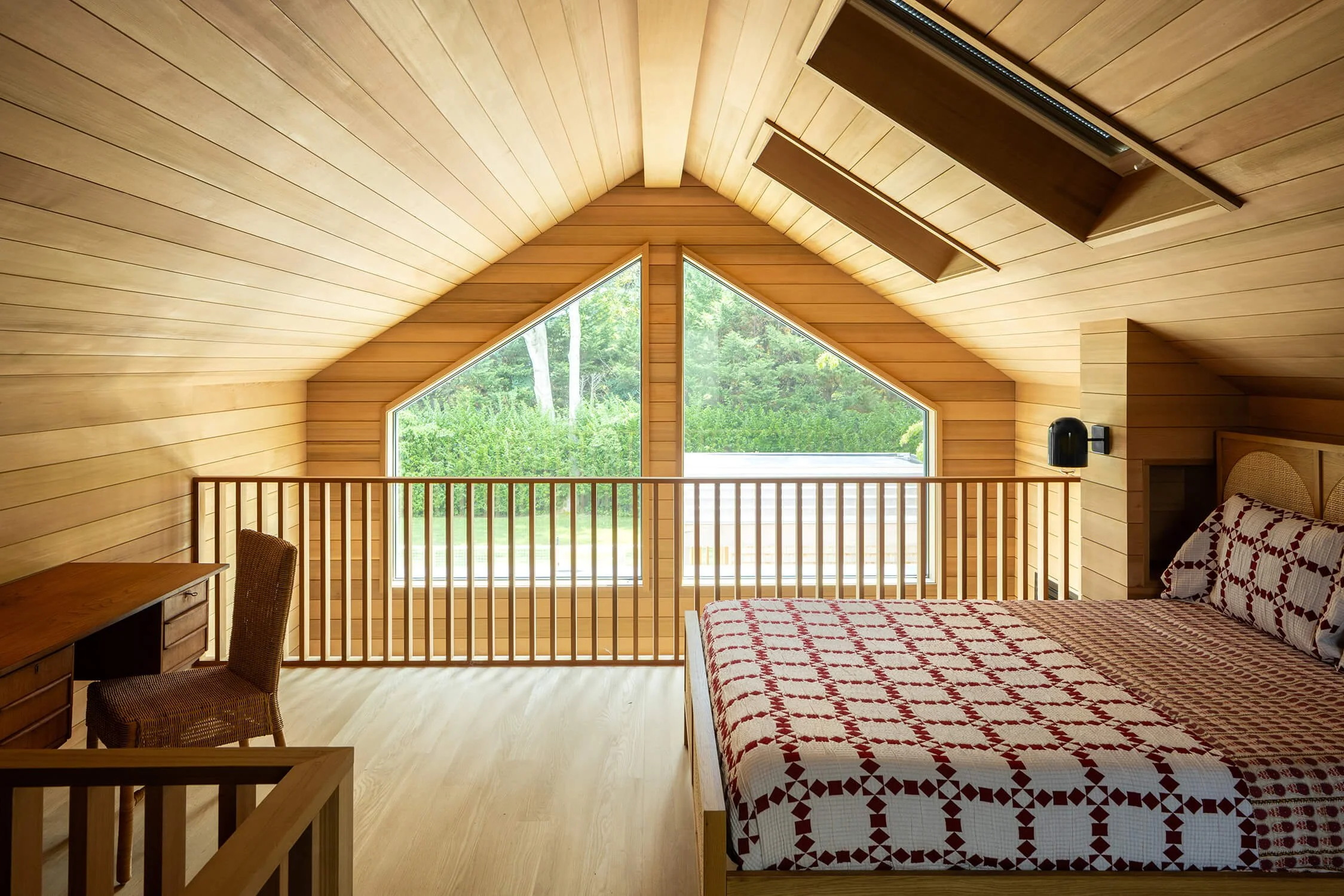 Pool house loft with vaulted cedar shiplap ceiling, triangular gable window overlooking garden, wood railing, and vintage quilt bedding, photographed by Brian Berkowitz Photography in the Hamptons