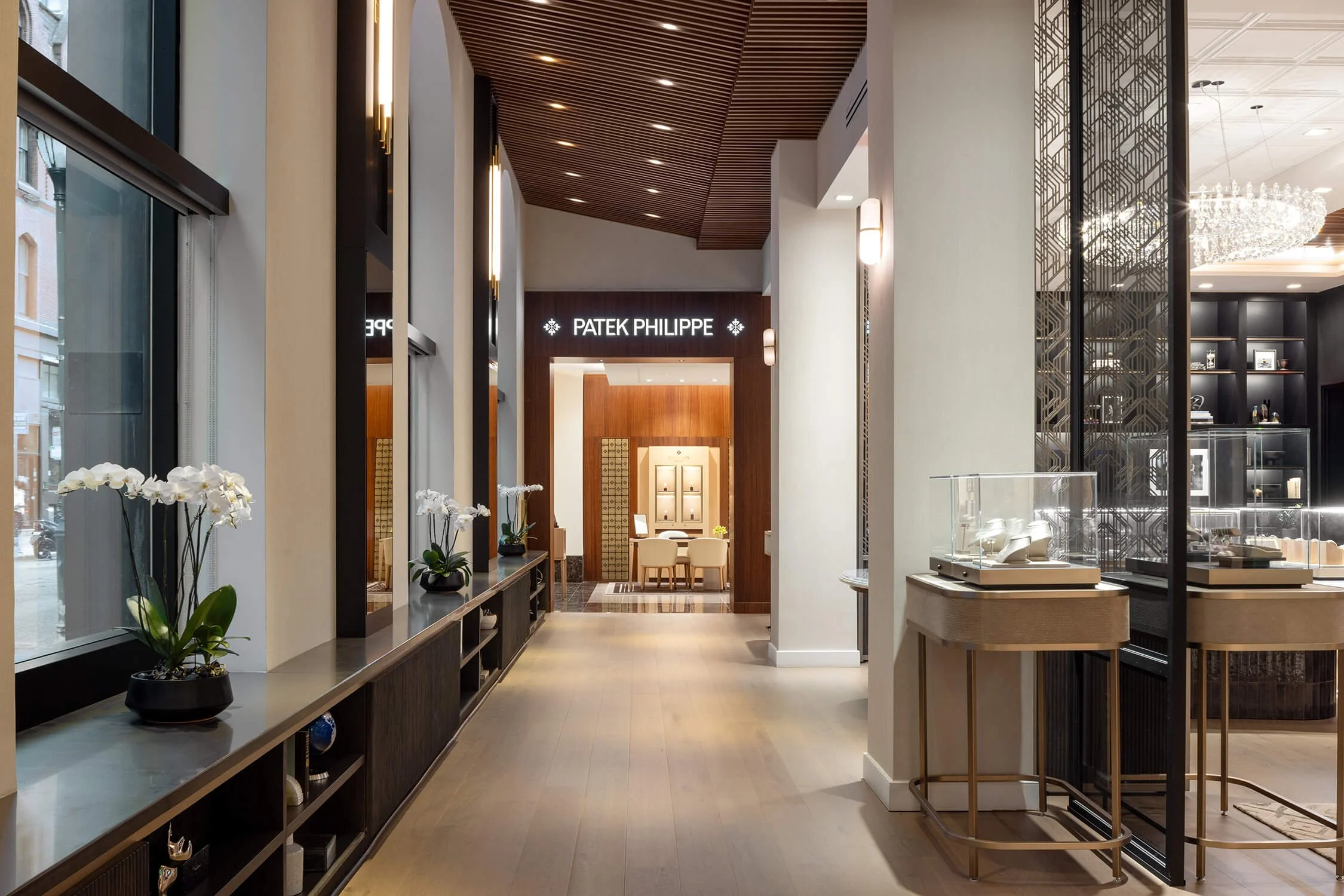 Refined retail corridor leading to a Patek Philippe boutique, featuring modern glass cases, decorative metal partitions, and bright natural light from large windows