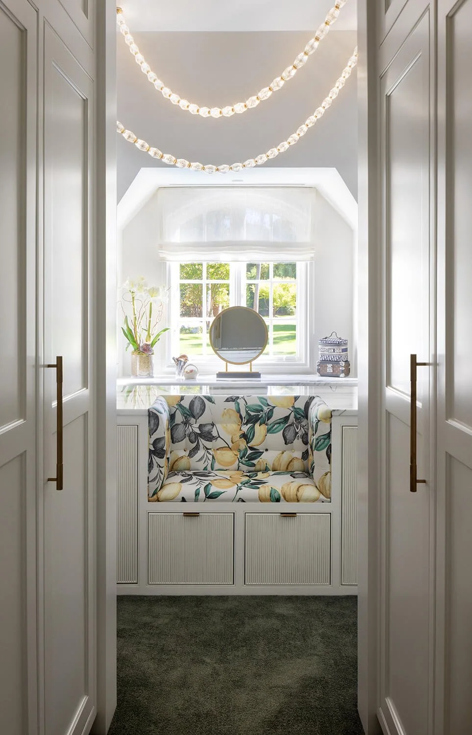 dressing room nook with pearl bead chandelier, floral bench, marble countertop, round brass mirror, and floor-to-ceiling closet doors, photographed by Brian Berkowitz Photography on Long Island