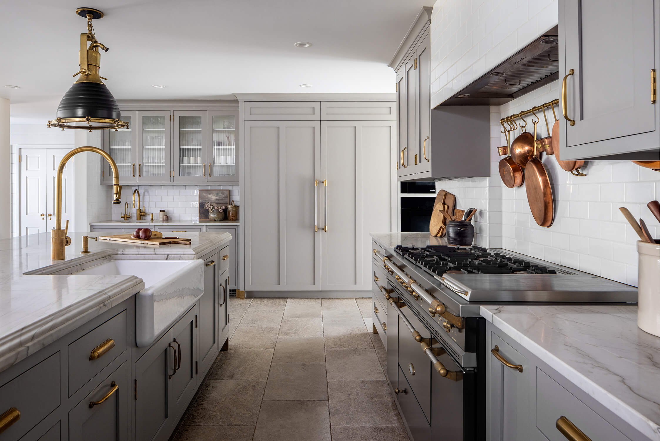 Chef's kitchen with gray shaker cabinetry, brass hardware and faucet, marble farmhouse sink, copper pot rack, and stone tile floor, photographed by Brian Berkowitz Photography on Long Island