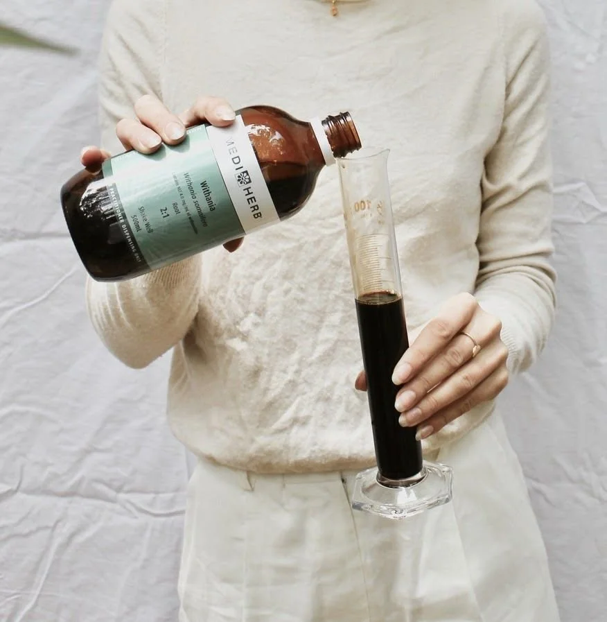 Person pouring a dark liquid from a brown bottle into a tall graduated cylinder.