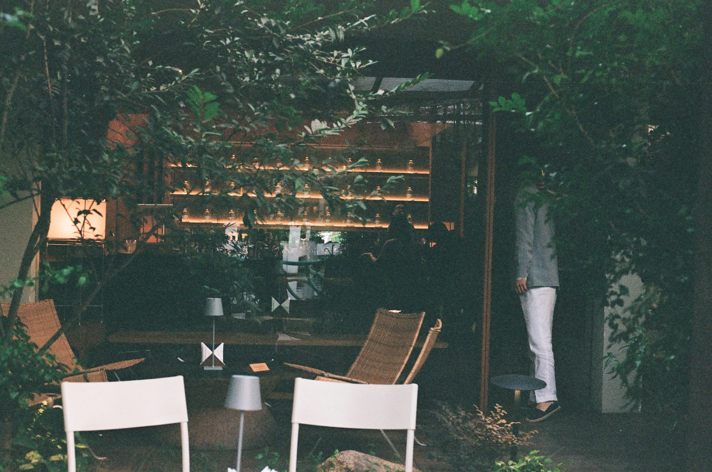 A dimly lit outdoor area with green plants and trees, a bar with bottles on shelves, white chairs, and a person standing with only their lower body visible.