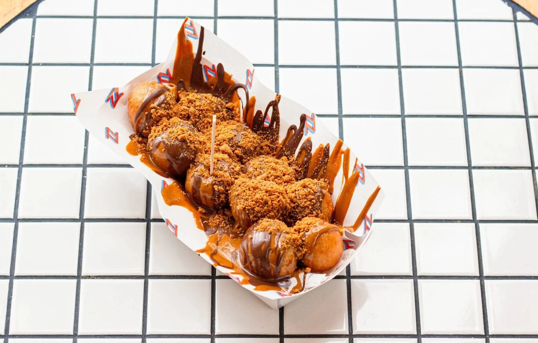 Deep-fried candy bars topped with chocolate drizzle and crushed nuts, served in a paper boat on a tiled surface.