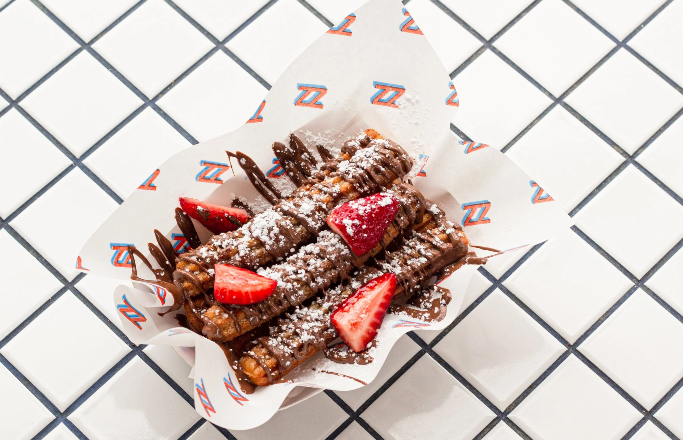 Chocolate waffles topped with strawberries and powdered sugar, served on branded paper with a checkered surface underneath.