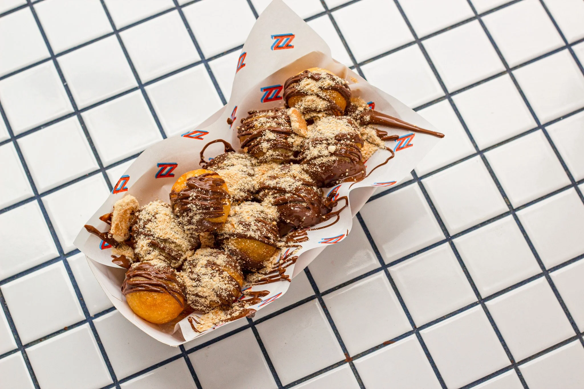 A paper food tray with three cannoli topped with chocolate drizzle, crushed nuts, and cookie pieces, placed on a white tile surface with black grout lines.