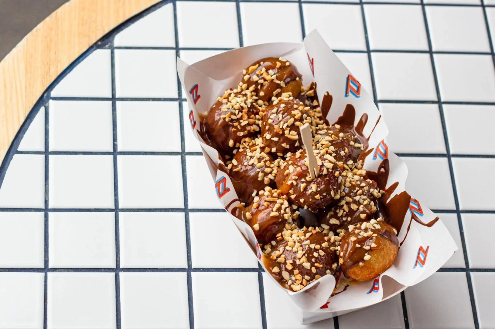Deep-fried donuts topped with chopped nuts and drizzled with chocolate sauce served in a paper boat