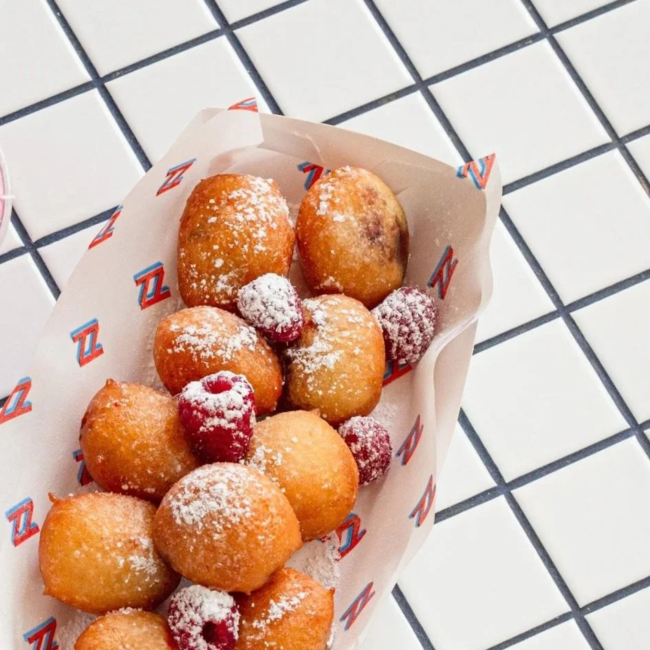 A paper-lined basket with fried dough balls dusted with powdered sugar and fresh raspberries on a white tiled surface.