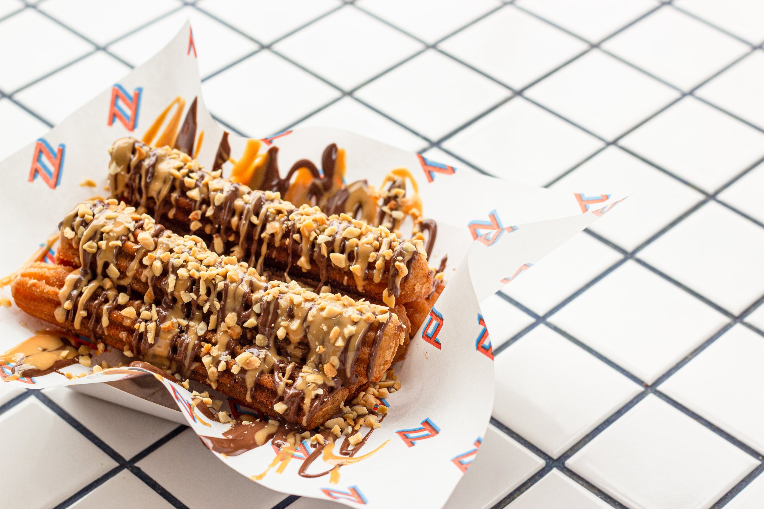 Close-up of three fried dough pastries topped with chocolate, caramel, and chopped nuts, wrapped in paper with a red and blue design, sitting on a black and white checkered surface.