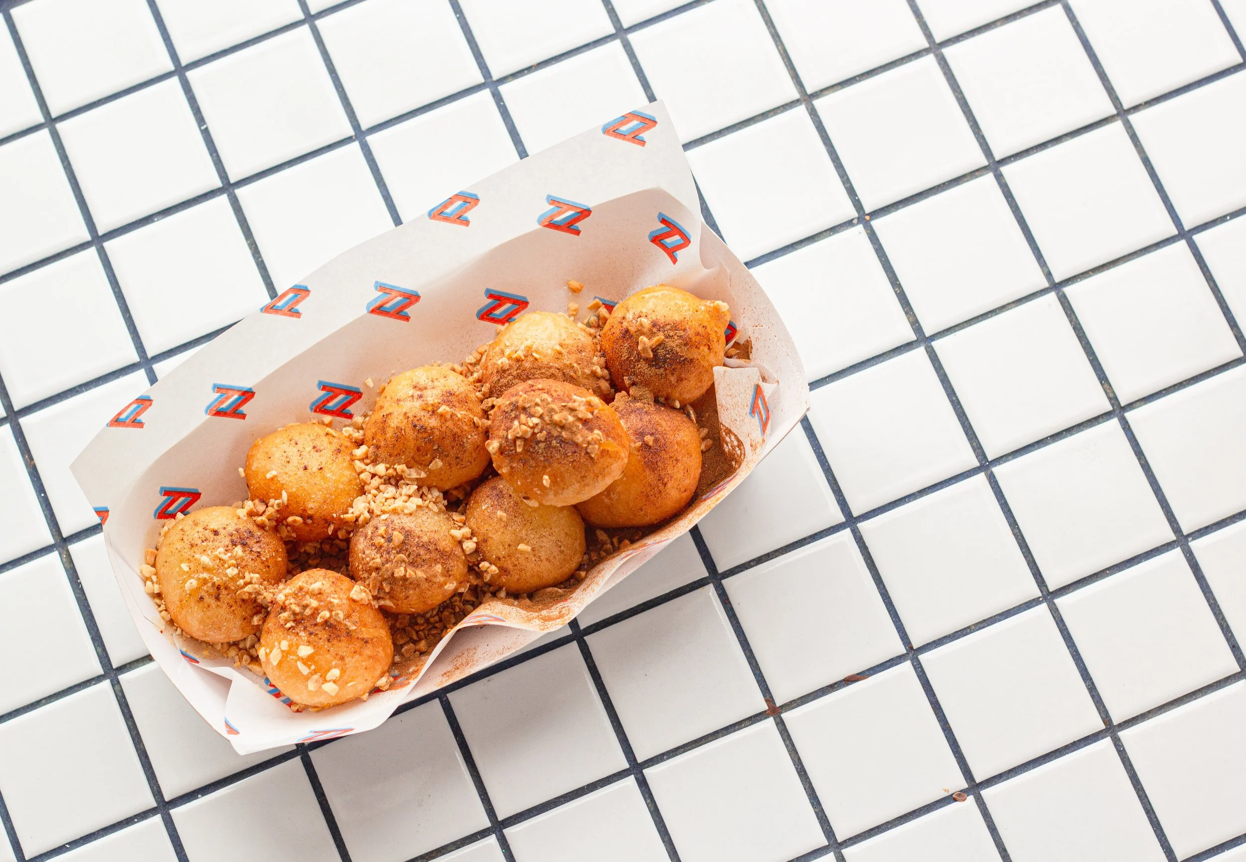 A paper tray filled with small, round, golden-brown fried balls, garnished with chopped nuts, resting on a white tiled surface with black grout lines.