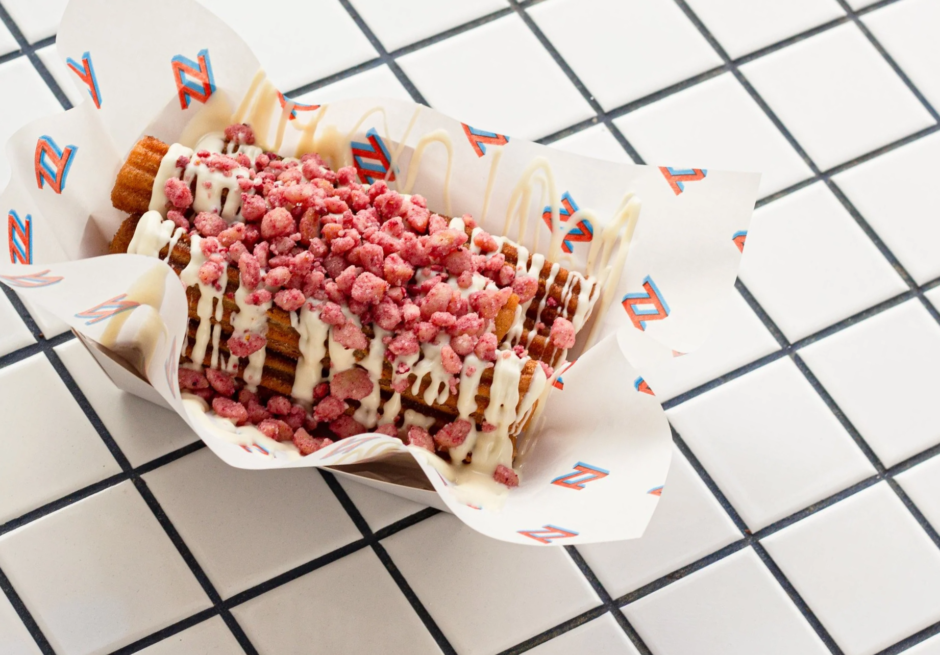 A waffle topped with white chocolate drizzle and pink crushed candy pieces, placed in a paper holder on a white tiled surface.