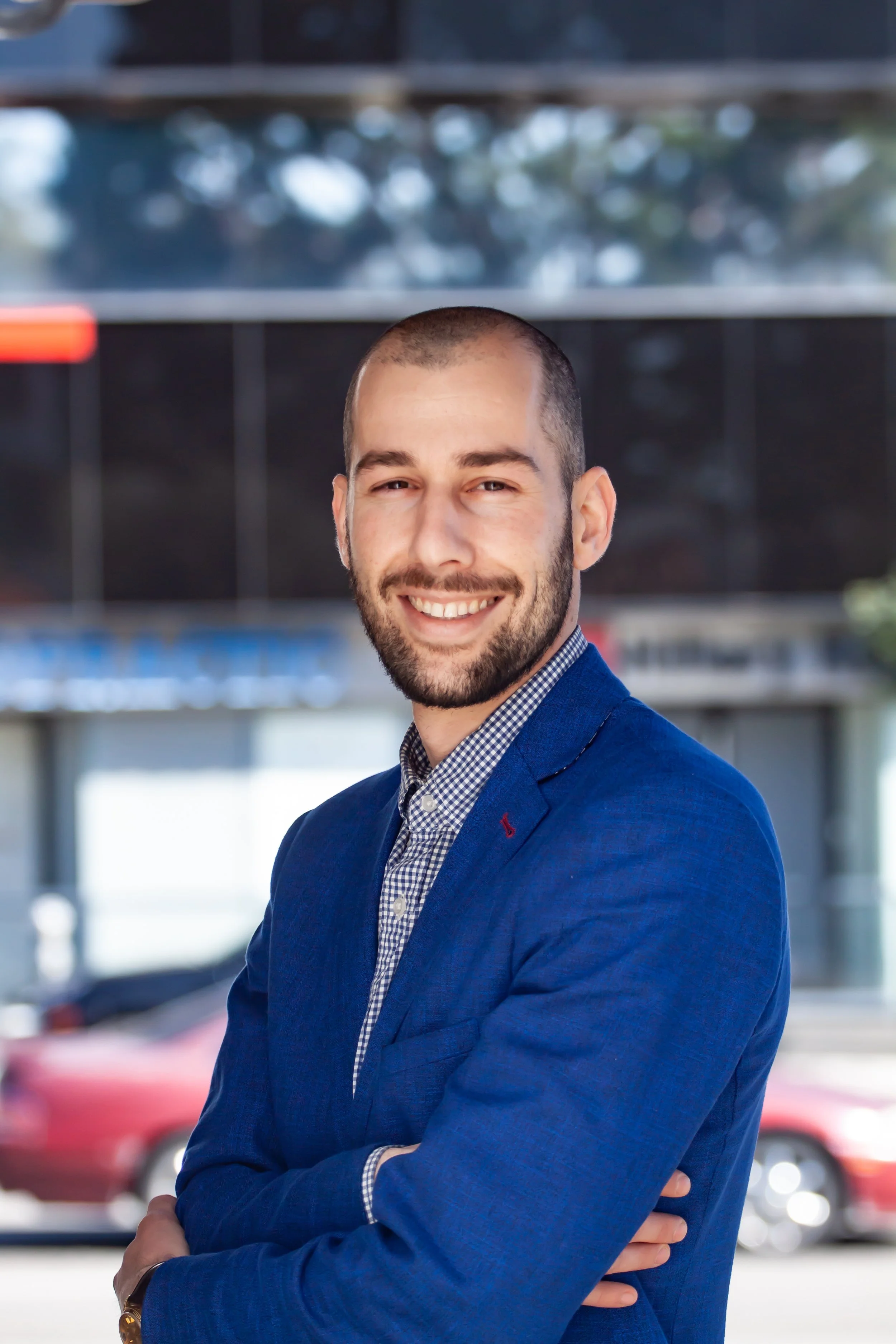 A man with a beard and short hair, wearing a blue blazer and checkered shirt, smiling with arms crossed, standing outdoors in front of buildings and trees.