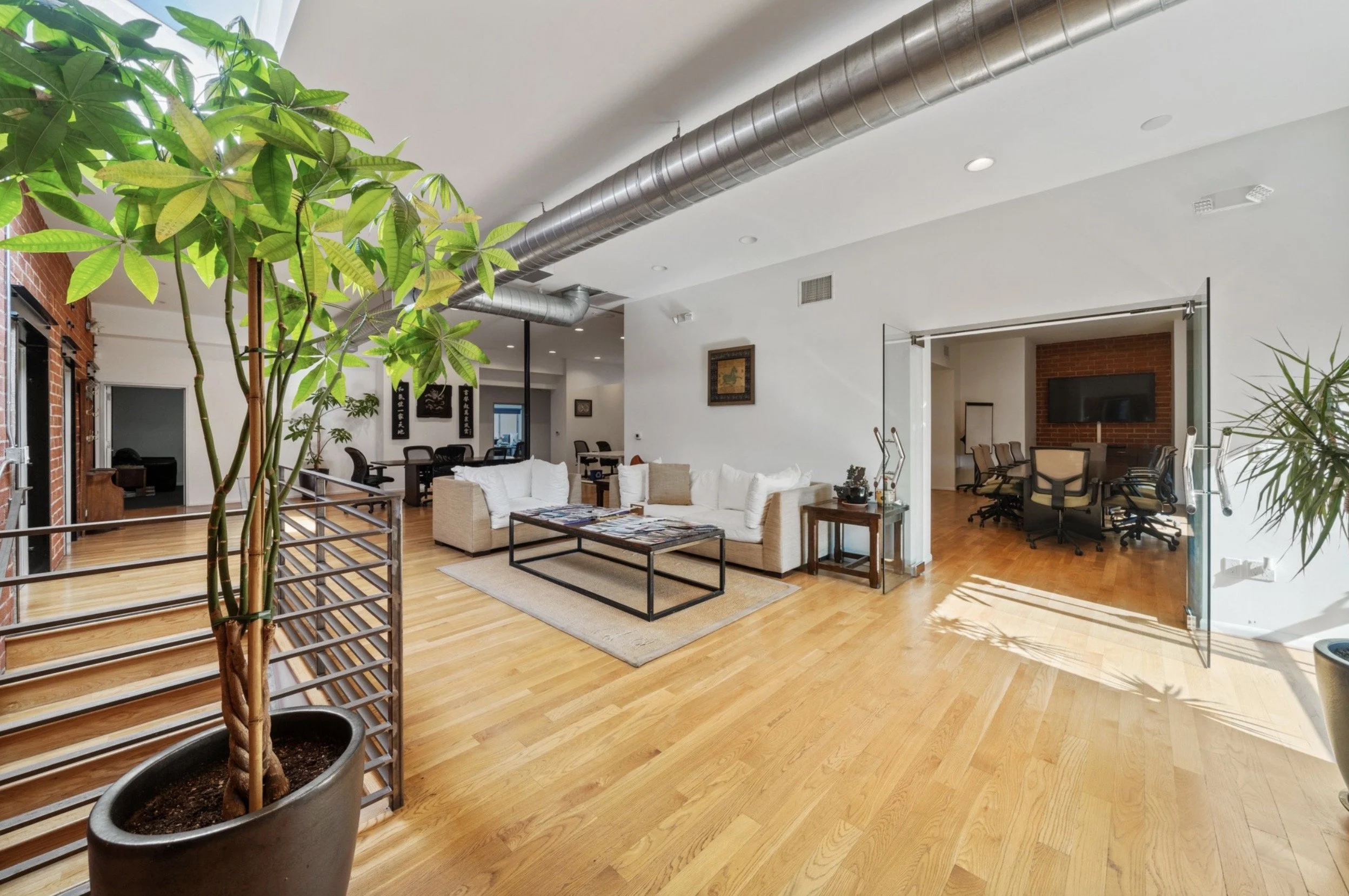 Bright modern office or co-working space with white sofas, side tables, a dining table with chairs, and glass sliding doors leading to a conference room with a large TV, wooden floors, potted plants, and exposed ventilation ductwork.
