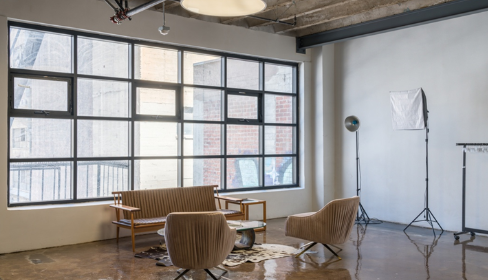Interior of a modern studio with large windows, a brown sofa, two matching armchairs, a floor lamp, and light setup, with a concrete ceiling and wooden floor.