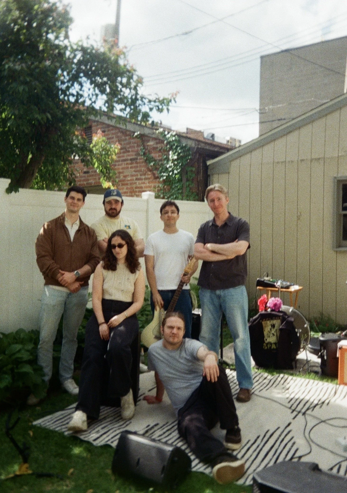 Group of six people standing and sitting outside in a backyard with a white fence, a tree, a brick house, and a gray building in the background. The group includes males and females, with some holding musical instruments, and there is a small table with flowers and musical equipment nearby.