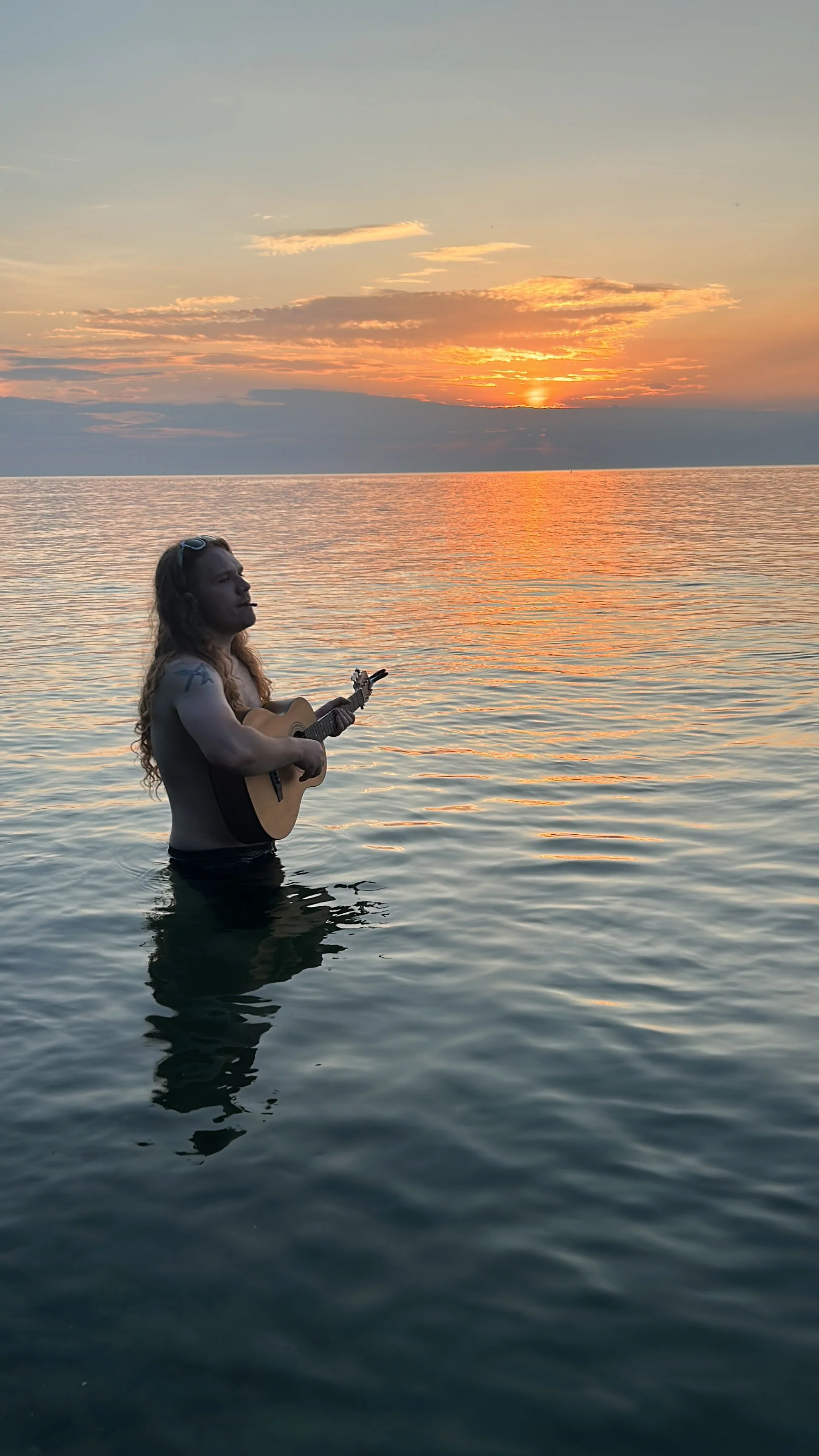 A person with long hair playing an acoustic guitar while standing in the water during a sunset. The sky is orange and pink, reflecting on the calm water surface.
