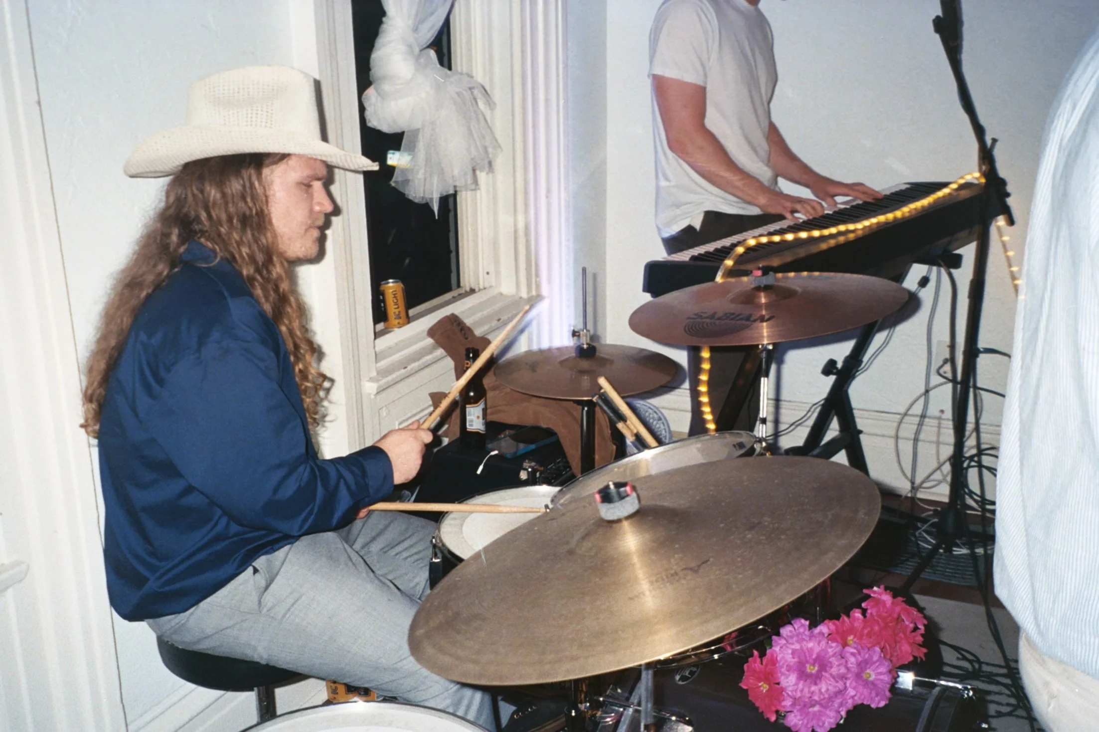 A man with long, curly hair and a cowboy hat playing drums, and another person wearing a gray shirt playing keyboard in a room with white walls and a window with curtains.