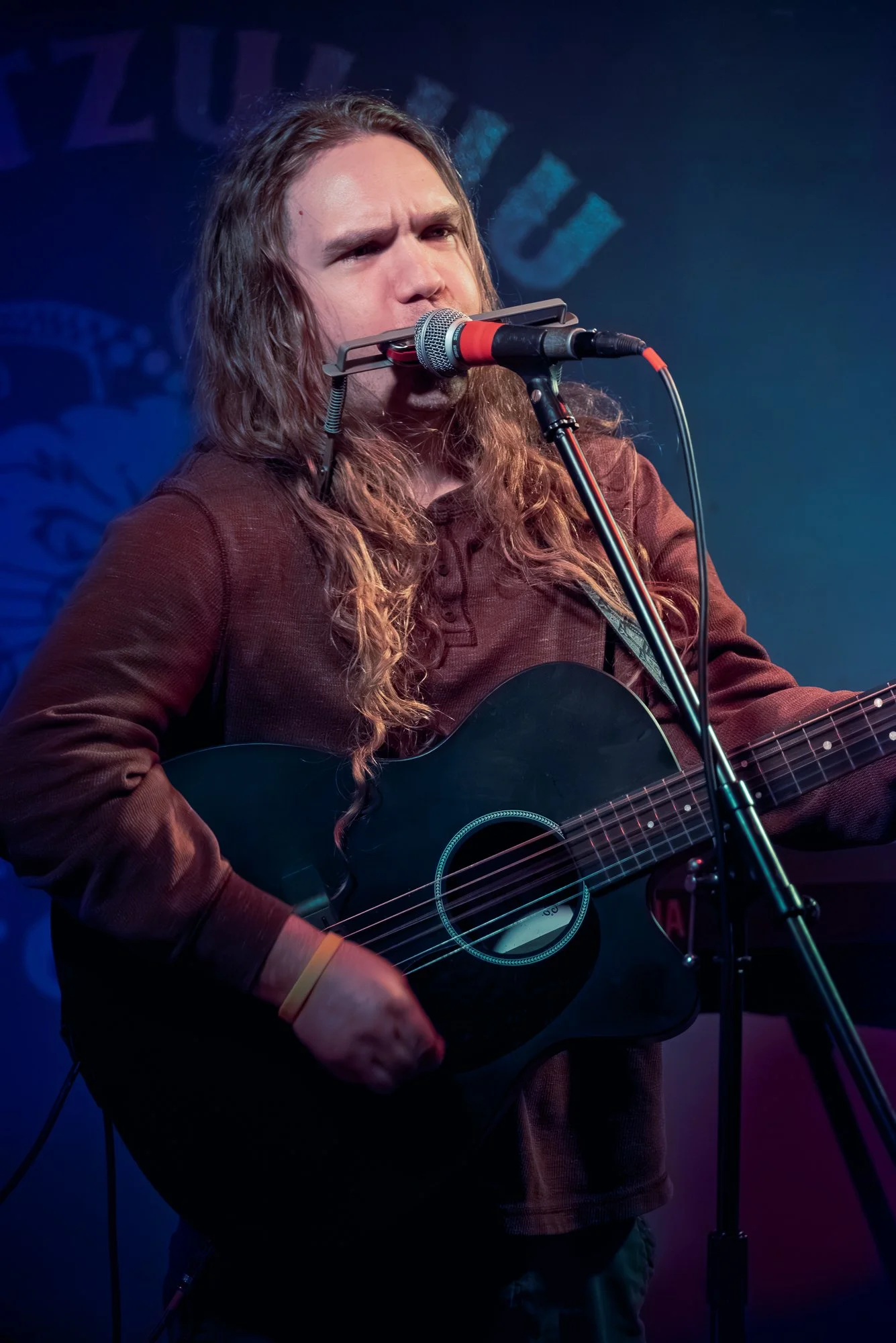 A man with long hair and a beard playing an acoustic guitar and singing into a microphone on stage with blue and purple lighting.