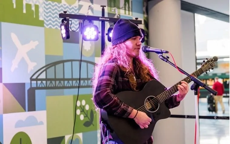 A young woman with long curly hair, wearing a dark beanie and plaid shirt, is playing an acoustic guitar and singing into a microphone on an indoor stage with purple lighting.