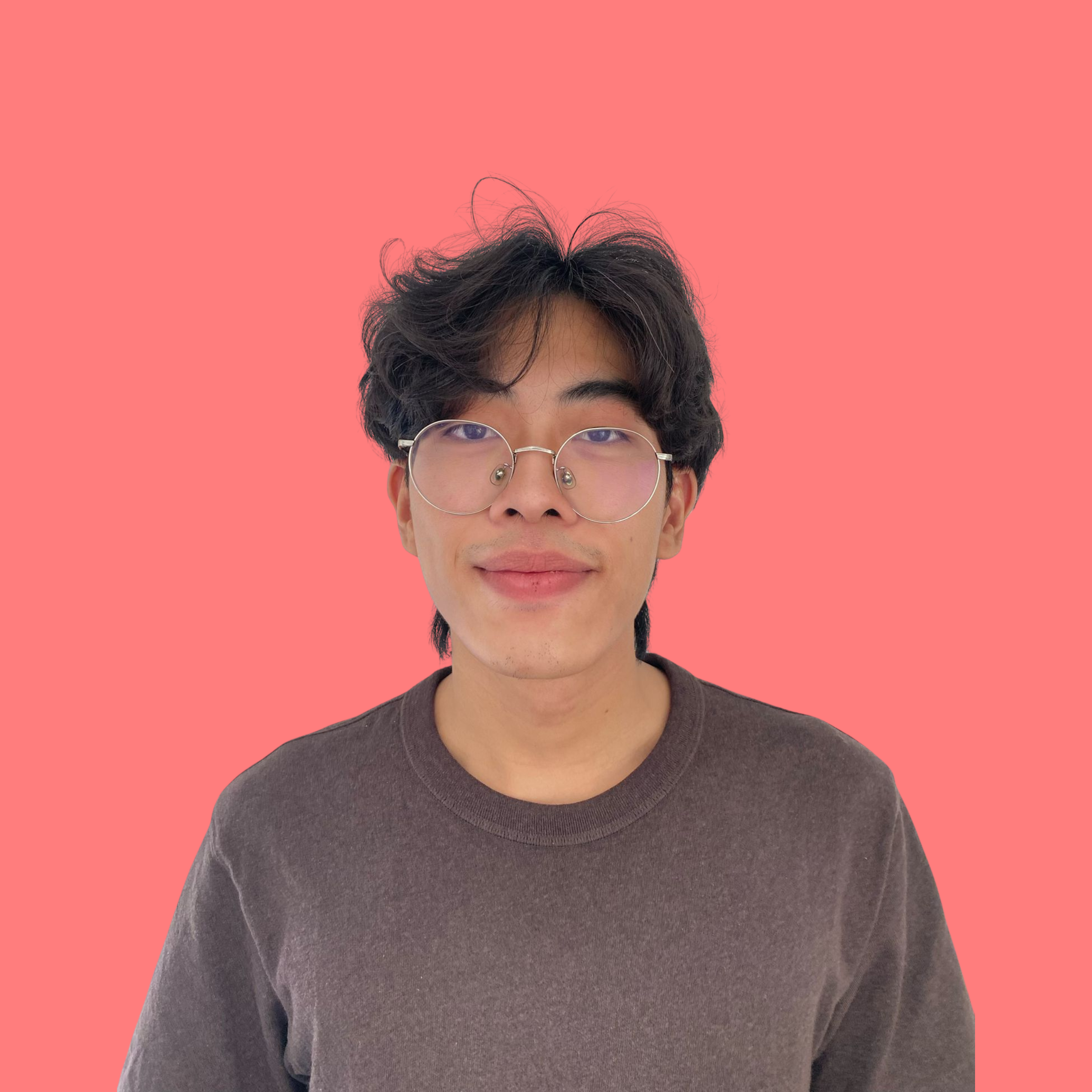 A young man with wavy dark hair, glasses, and light skin, wearing a brown sweater, standing against a pink background.