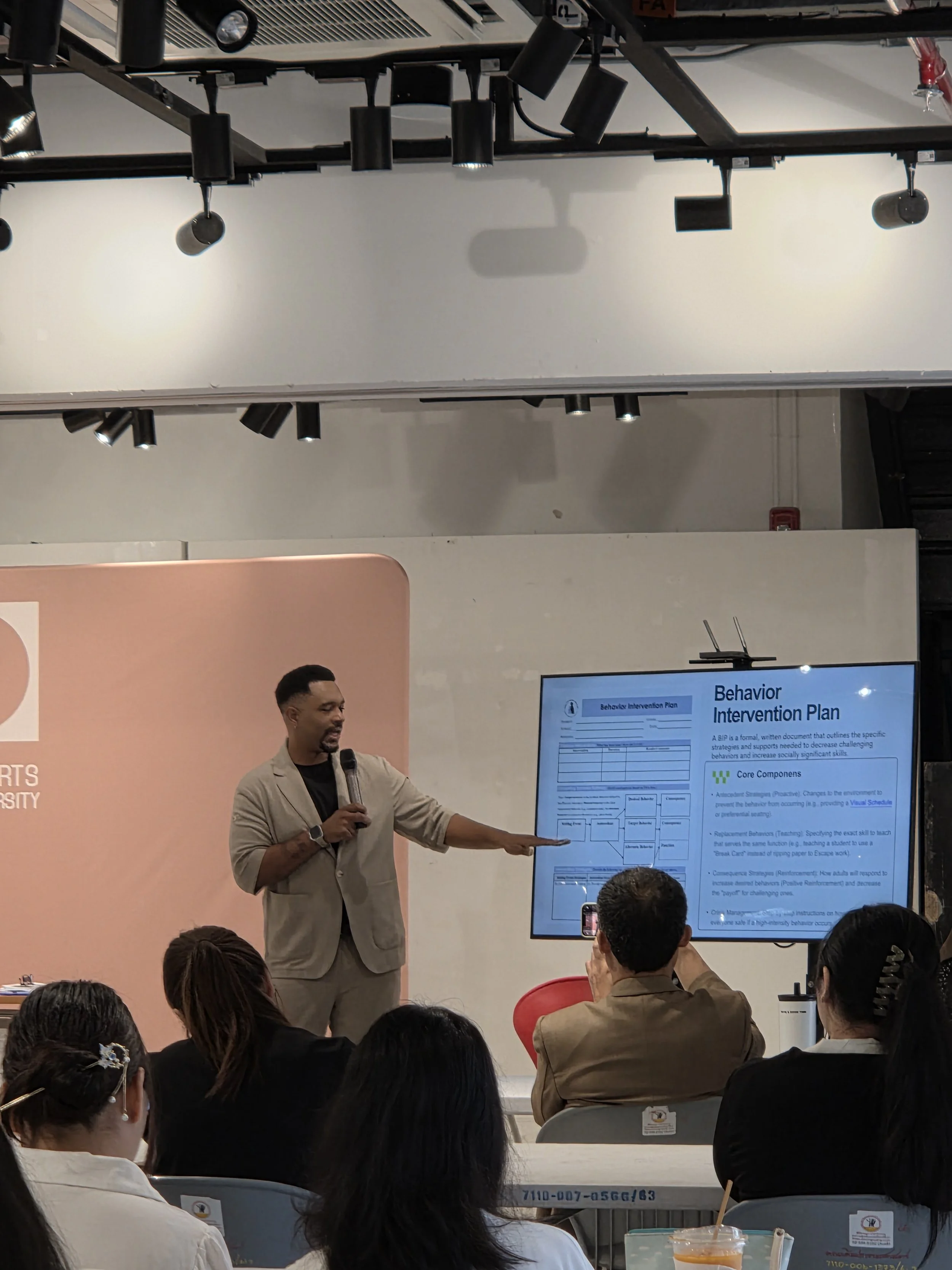 A speaker stands at the front of a modern conference room giving a presentation to an audience. He holds a microphone and gestures toward a screen displaying a slide titled “Behavior Intervention Plan.” Attendees seated in rows watch the presentation
