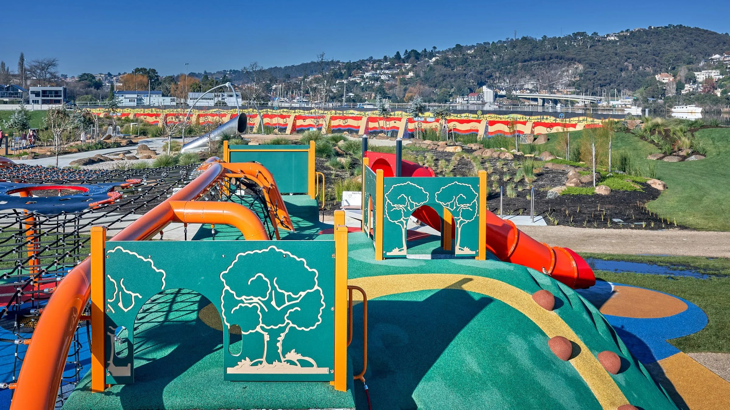 Colorful playground with slides, tunnels, and climbing structures on a grassy hill with a cityscape and hills in the background under a clear blue sky.