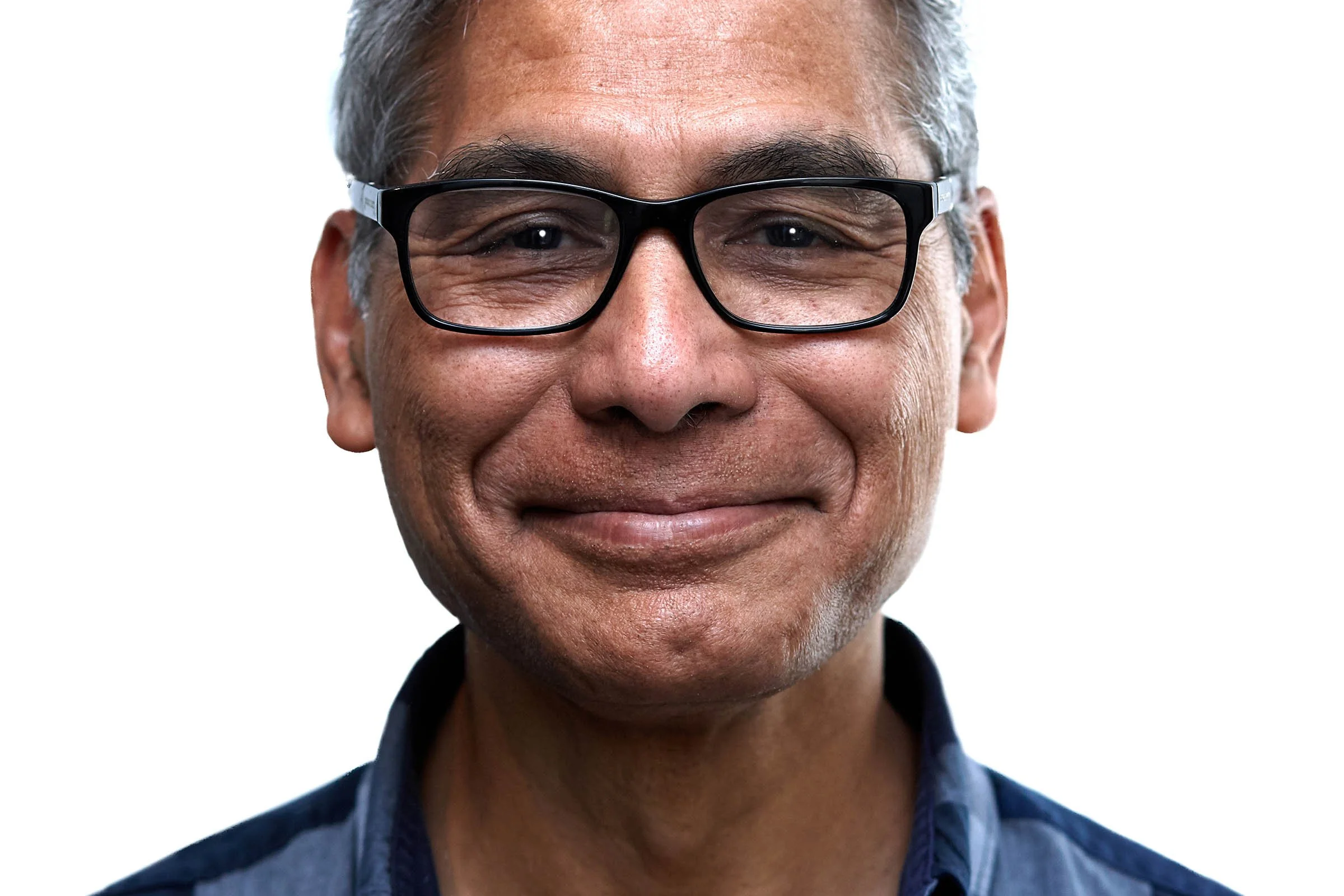 Close-up of a smiling middle-aged man with gray hair, wearing black-rimmed glasses and a dark blue shirt, against a white background.