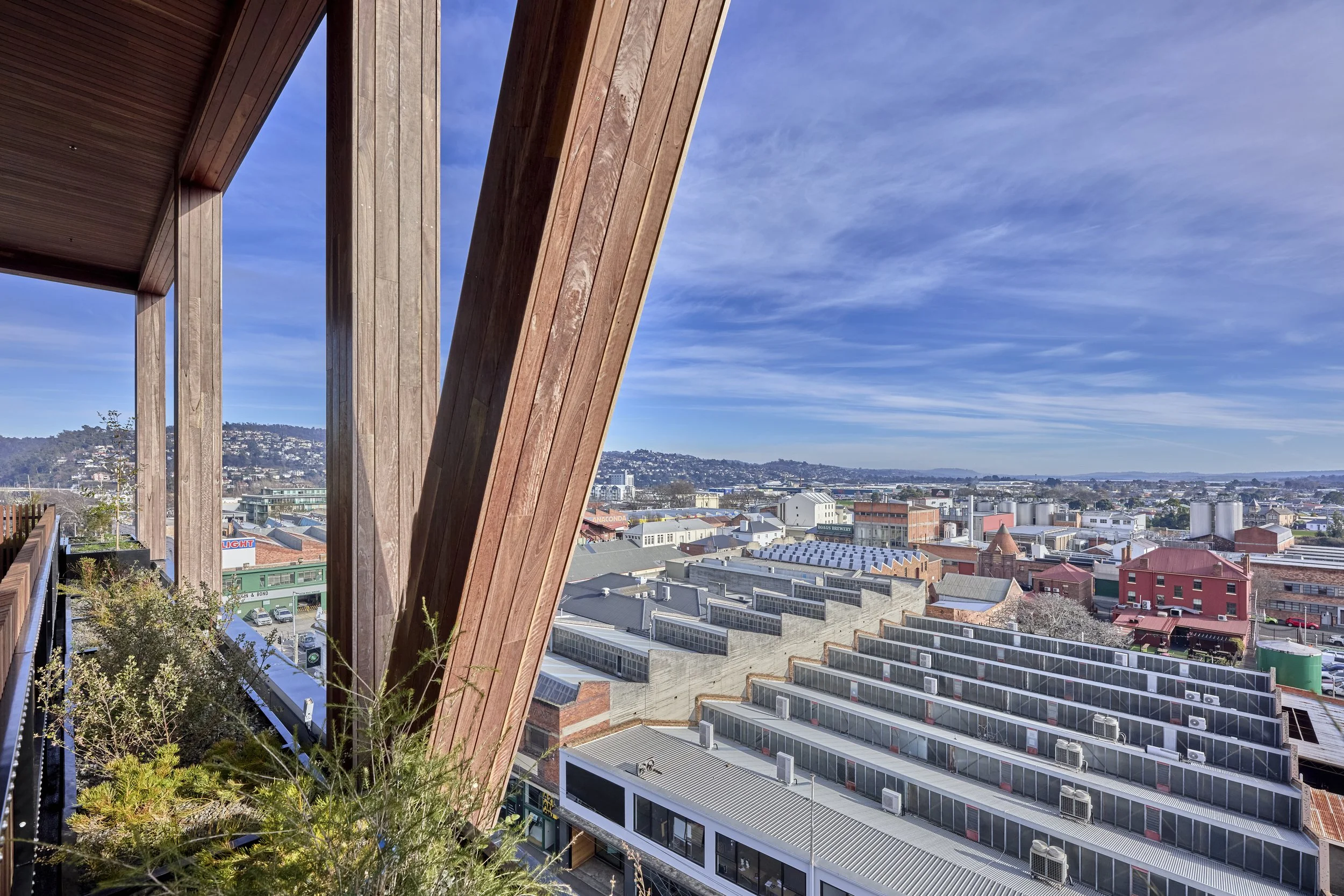 View of a cityscape from a balcony with wooden beams and plants in the foreground, under a partly cloudy sky.