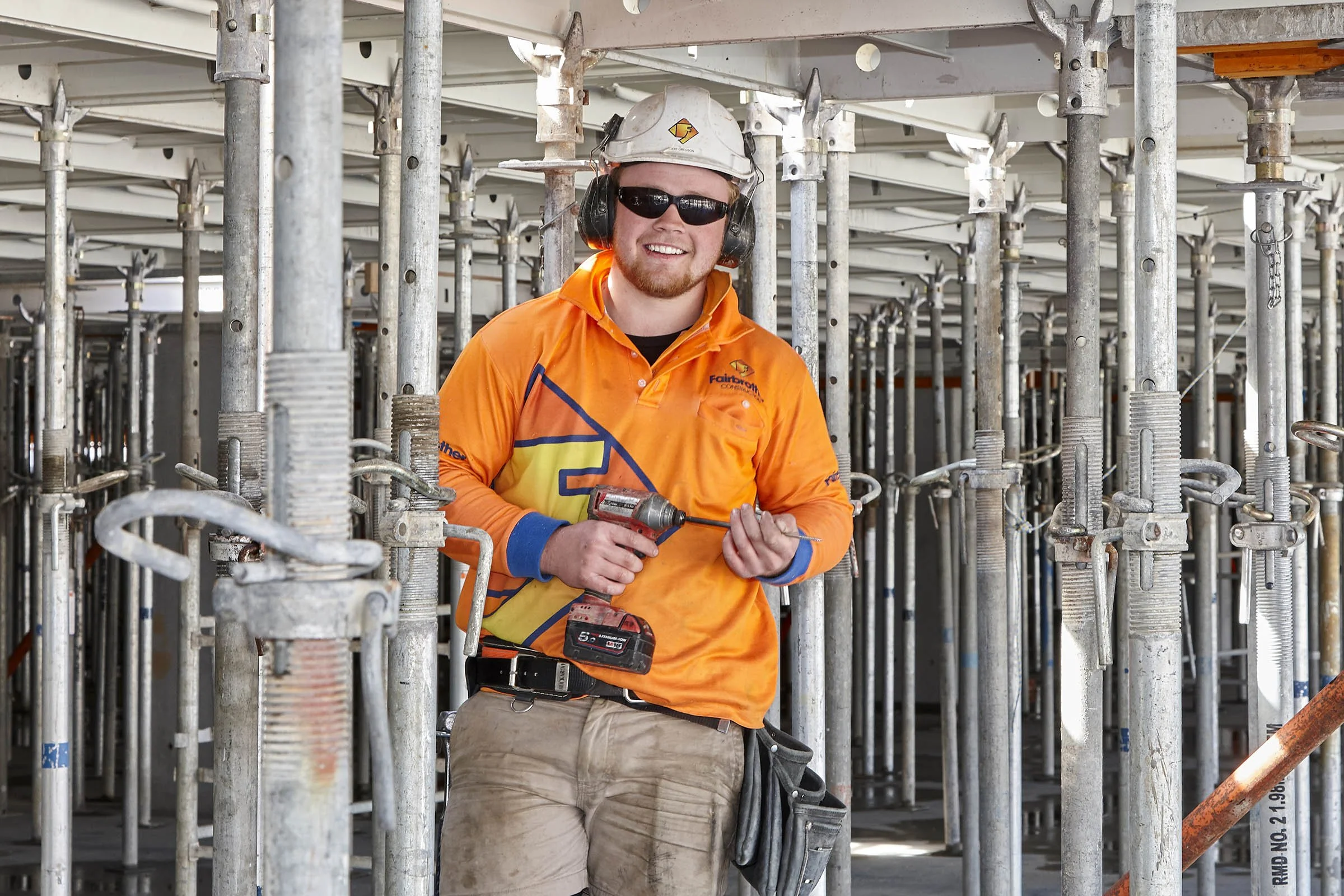 A construction worker in an orange shirt, wearing a helmet and sunglasses, is standing among scaffolding and holding a power drill.