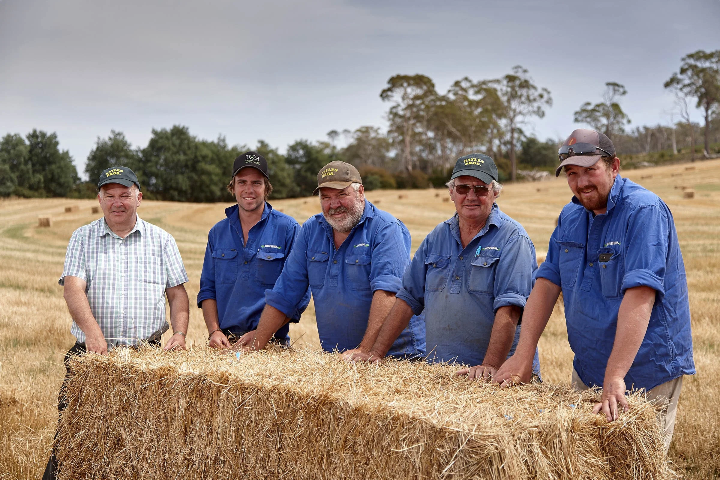Five men standing behind a hay bale in a farm field, wearing work shirts and hats.