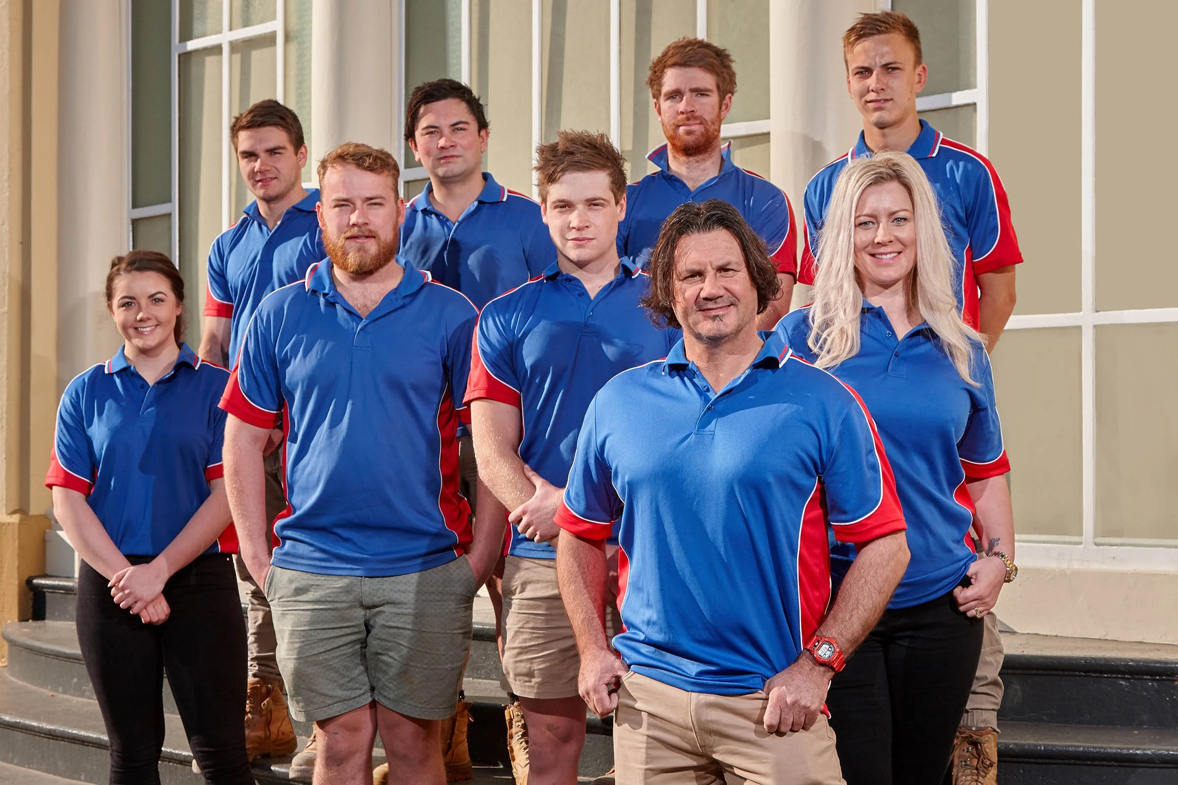 Group of nine people standing on a staircase outside, wearing blue sports shirts with red and white accents, posing for a team photo.