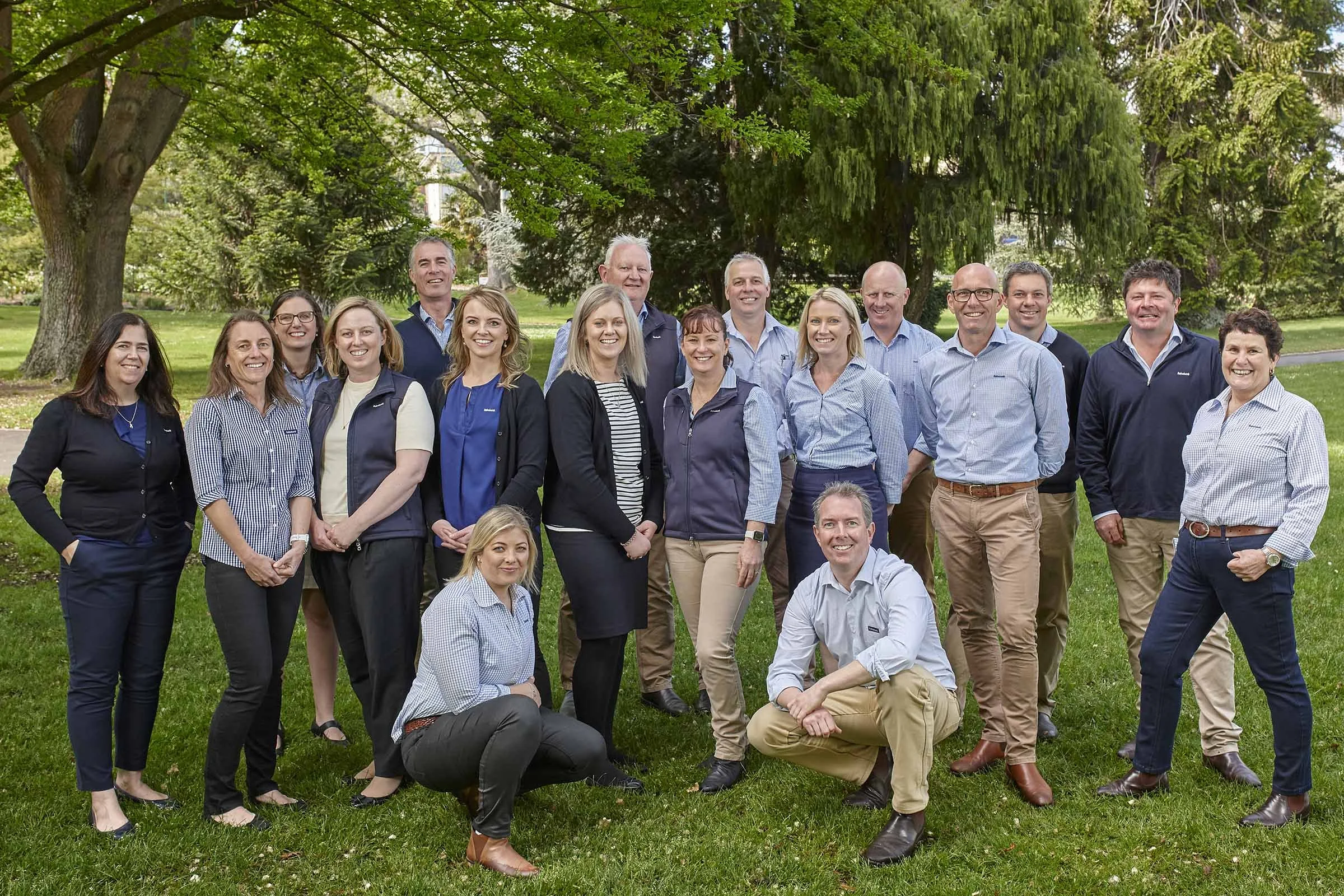 A group of 17 diverse adults, dressed in casual and business casual attire, standing outdoors on a grassy area with large trees in the background on a cloudy day.