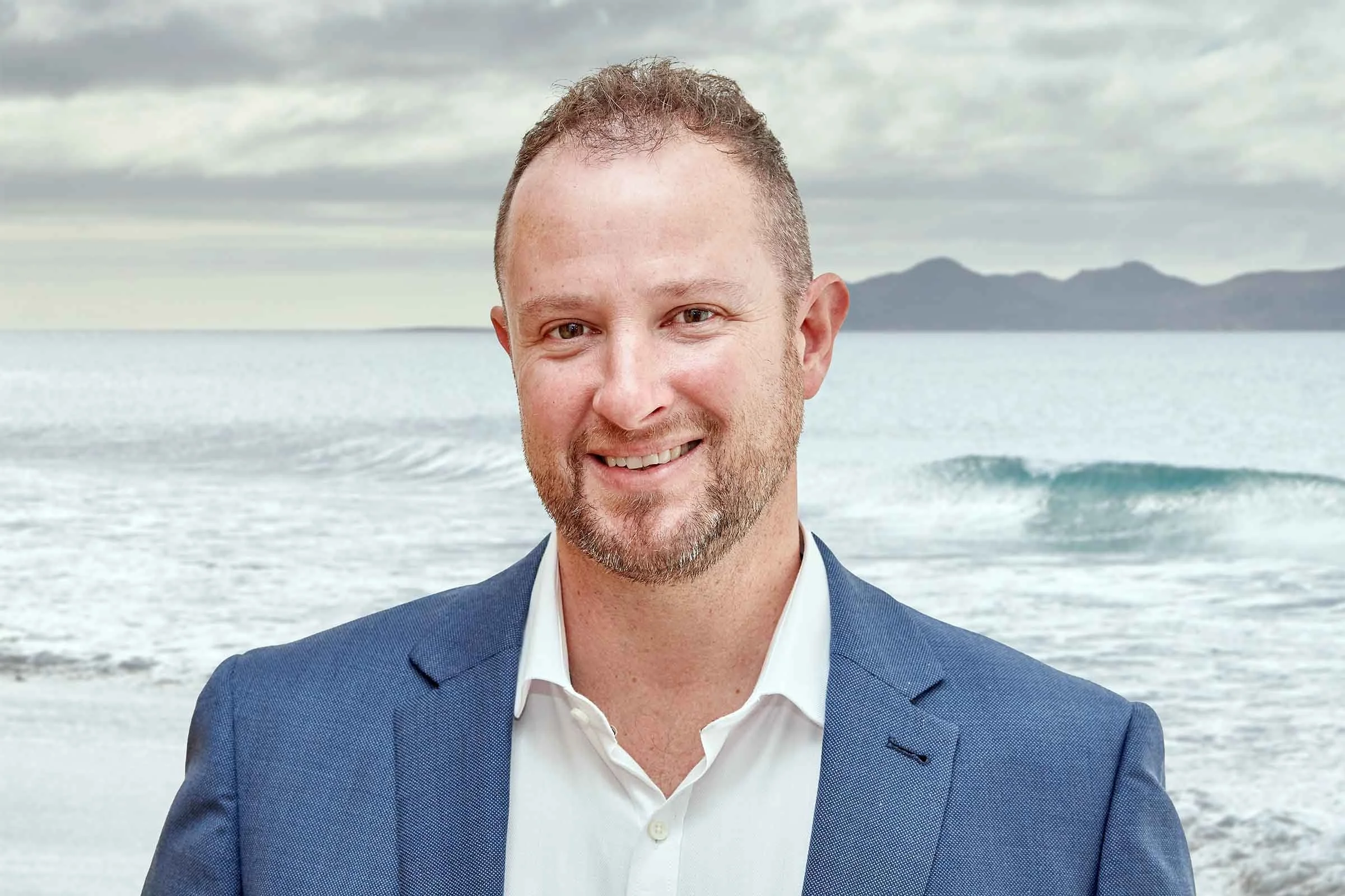 A man in a blue blazer and white shirt smiling at the camera with an ocean and mountains in the background.
