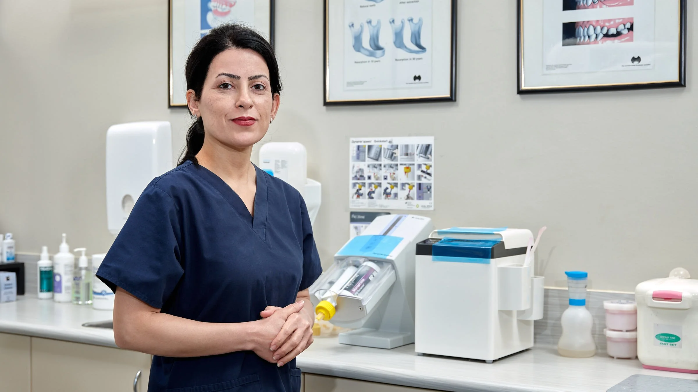 A woman in navy-blue scrubs standing in a medical or dental office with equipment and supplies on the counter behind her.