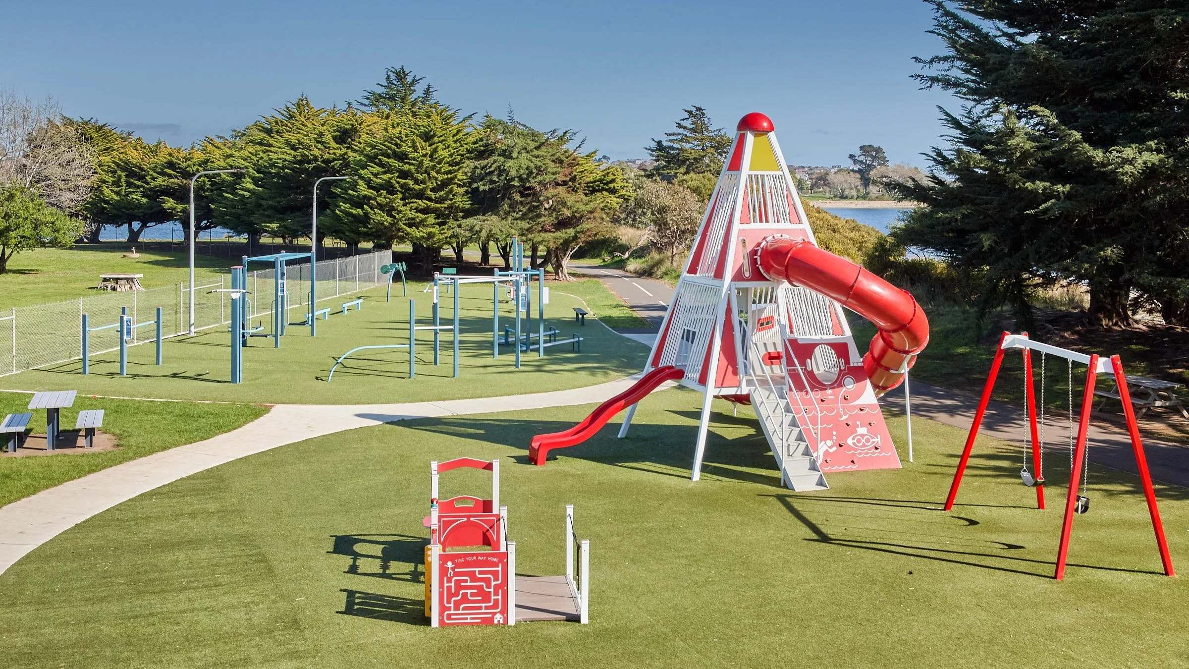 Children's outdoor playground with a large red and white play structure, slide, swings, and exercise equipment, set on green grass near trees and a body of water.