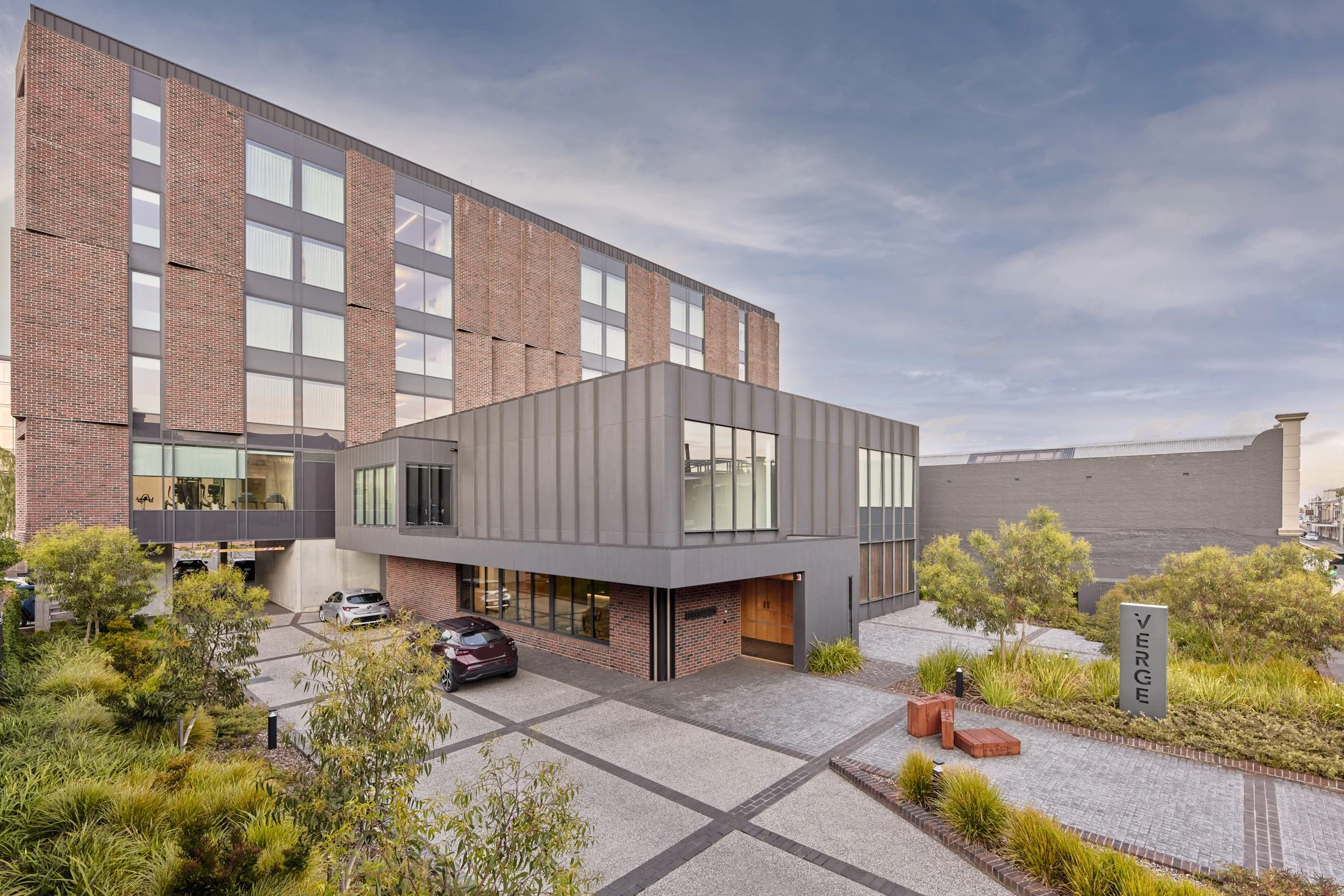 Modern multi-story building with brick and metal exterior, parking area, landscaped greenery, and a sign that says 'VIRGE'.