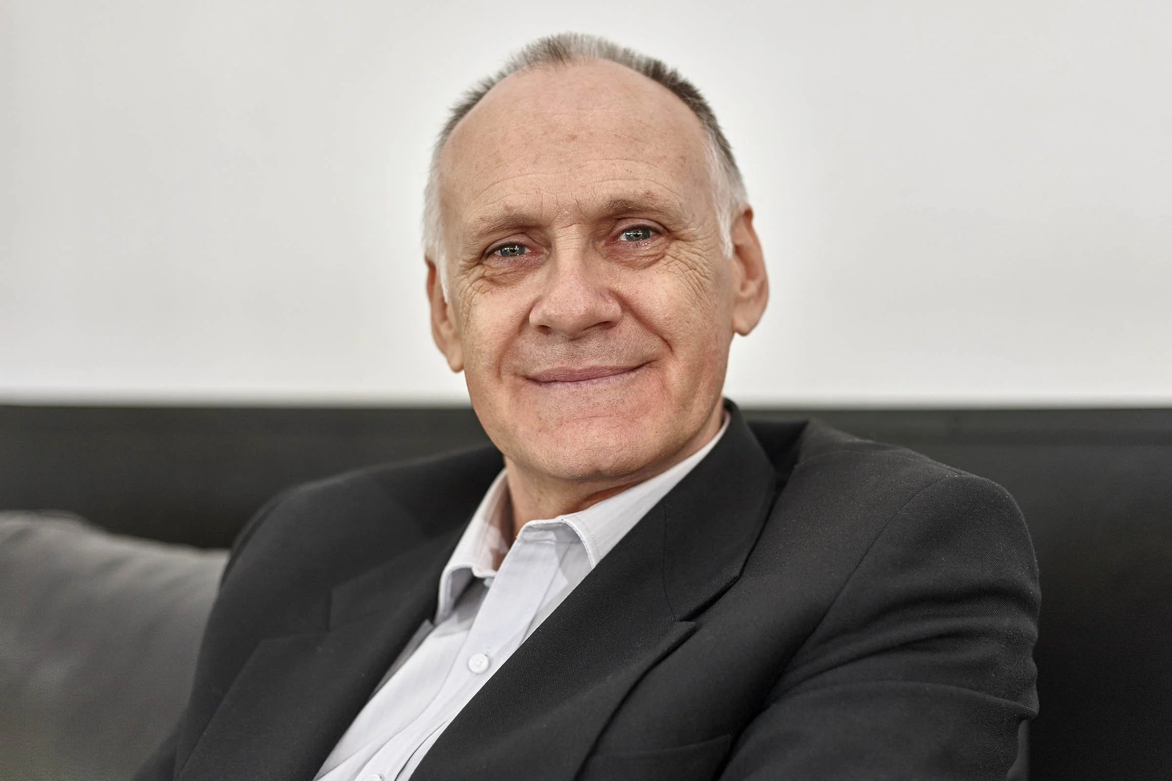A smiling middle-aged man wearing a black suit jacket and white shirt, sitting on a gray couch in front of a plain white wall.