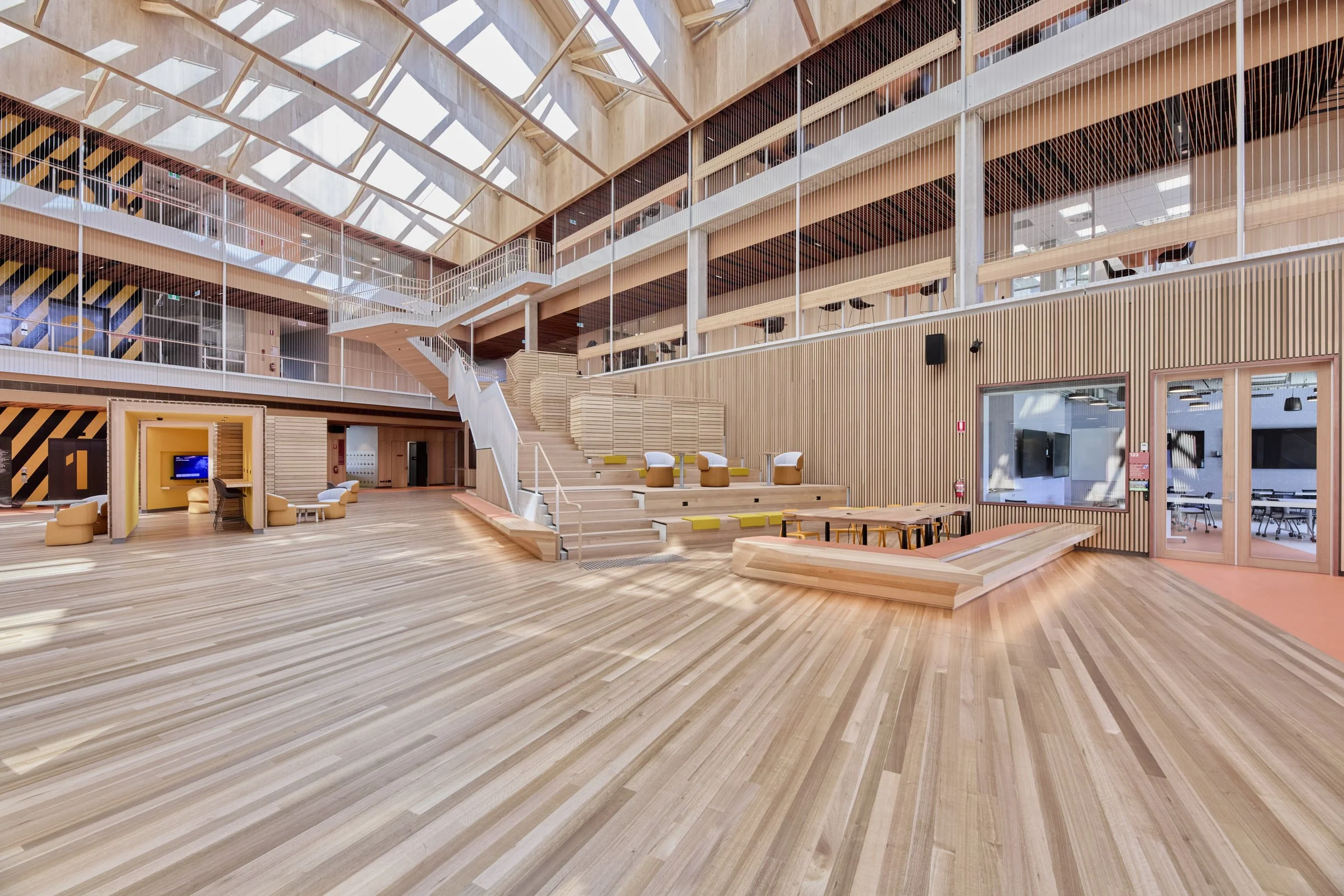 Modern interior with wooden floors and walls, open space with seating areas, stairs, and glass-walled rooms, under a large glass ceiling.
