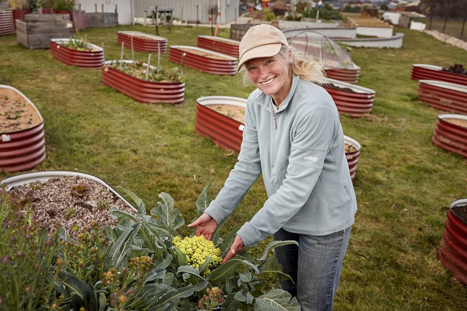 A woman in a light gray jacket and a beige cap is smiling while tending to a green plant in a garden with raised red metal garden beds in the background.