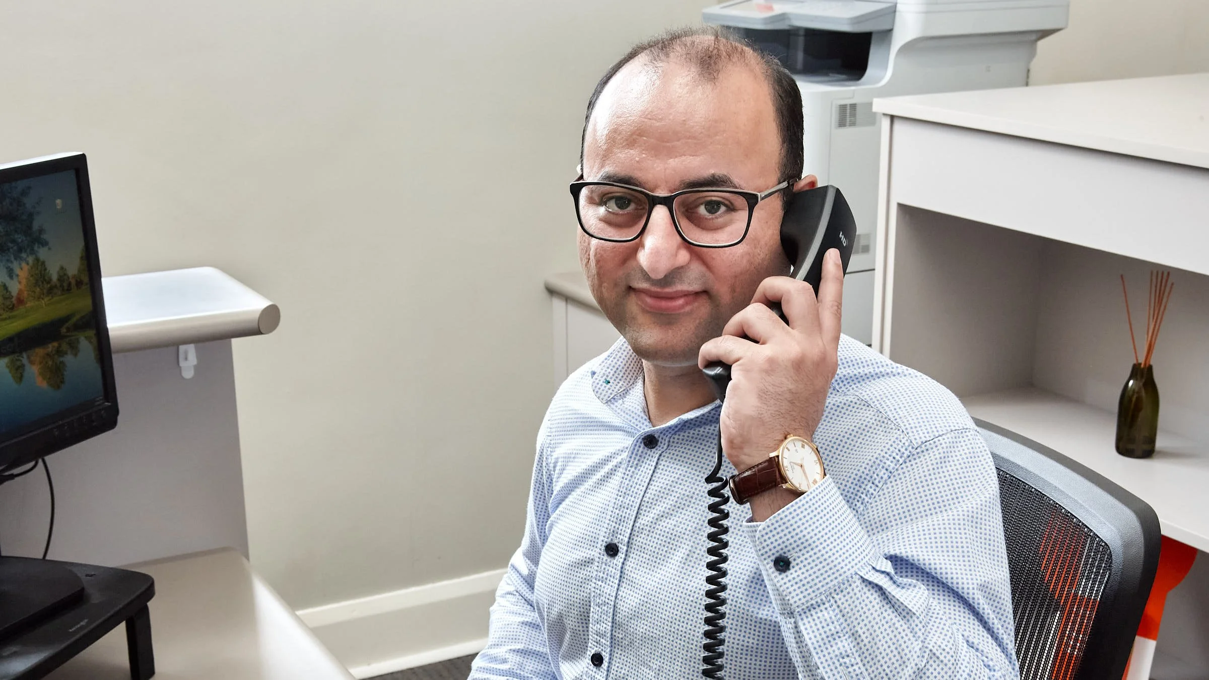 Man wearing glasses and a light blue dress shirt, sitting at a desk talking on a black corded telephone in an office. There is a computer monitor and a small white shelf nearby, with a white side cabinet and a decorative bottle with sticks in the bac