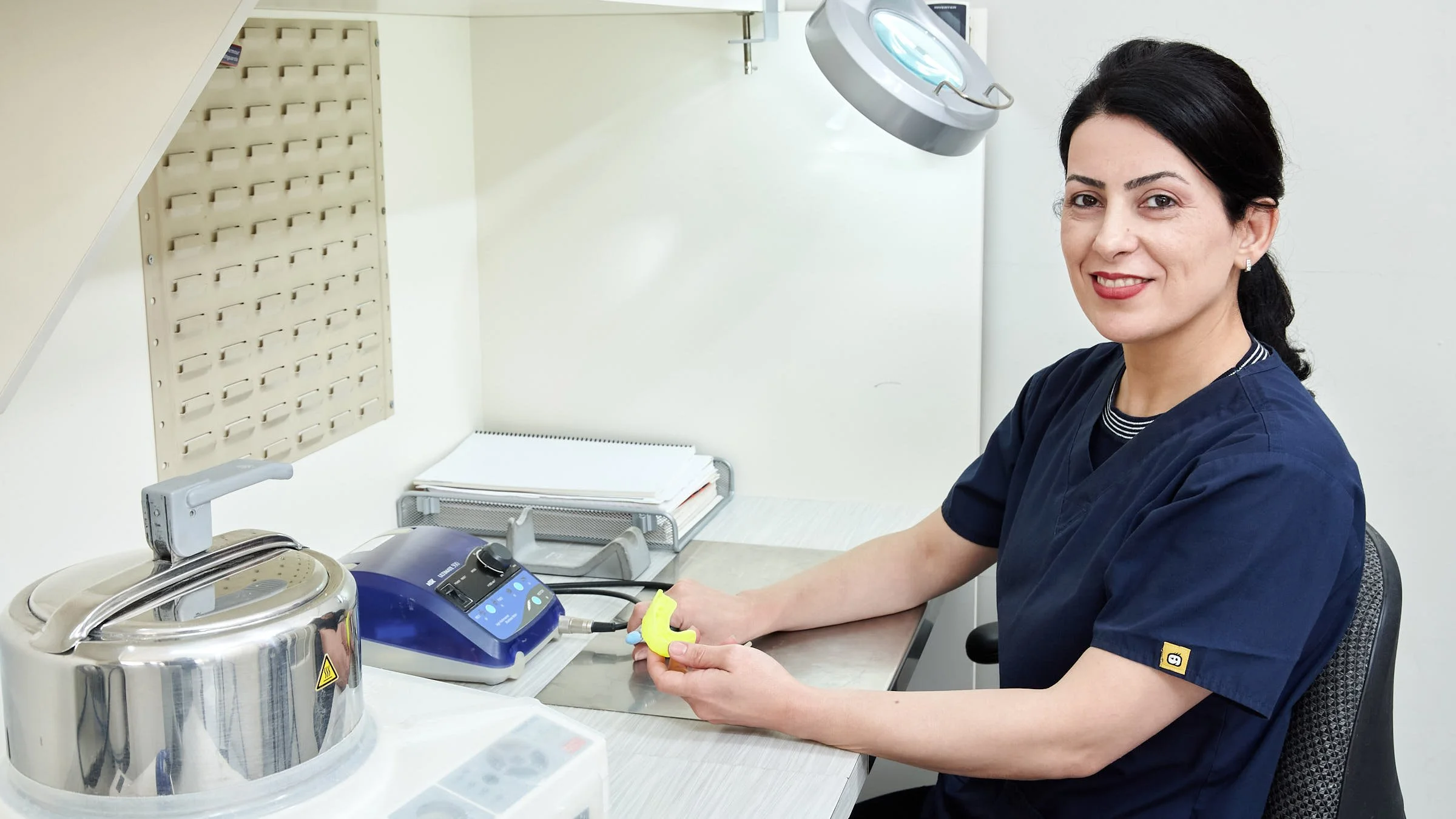 A female healthcare professional in navy scrubs sitting at a medical sterilizer machine in a clinic.