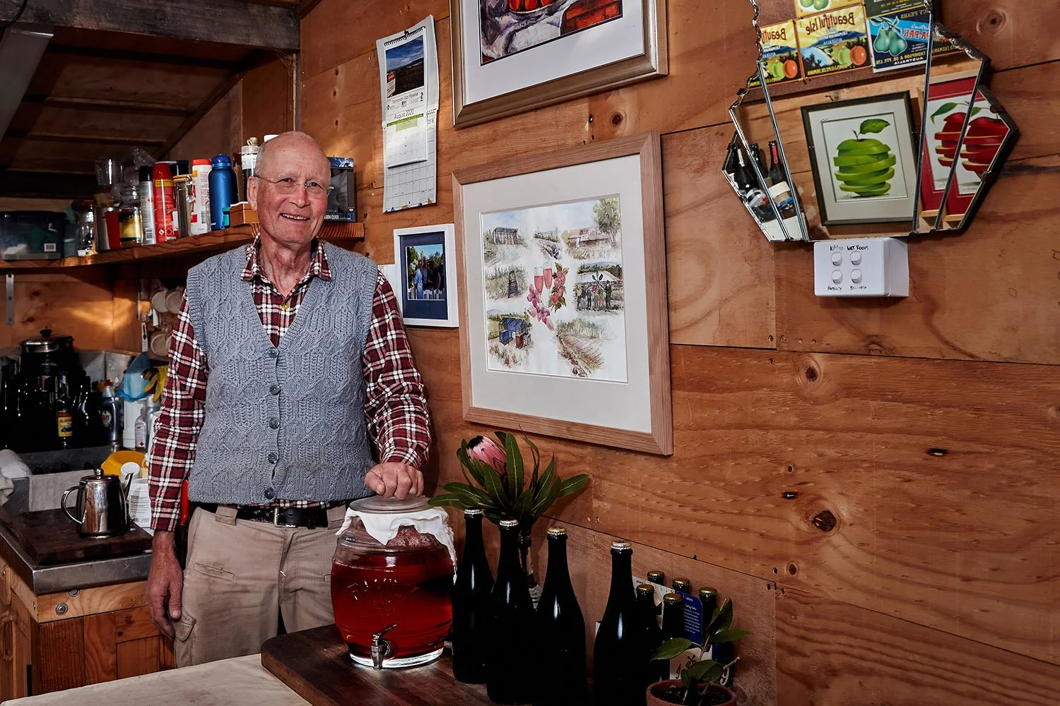 An elderly man with glasses, dressed in a plaid shirt and a knitted vest, standing in a cozy wooden kitchen. There are framed pictures and artwork on the wooden wall behind him, including a large watercolor and a garden-themed print. On the table in