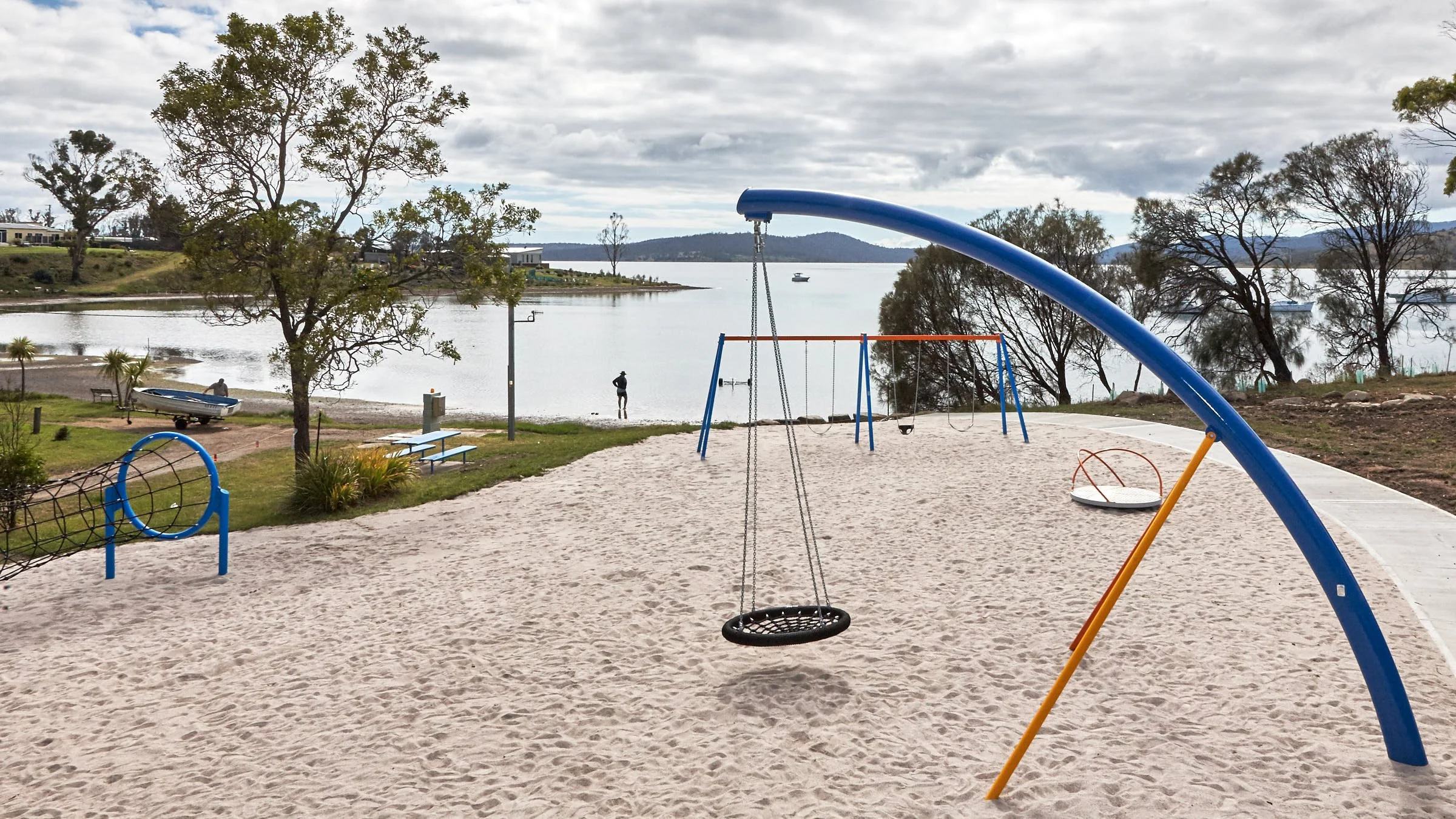 Playground with swings and climbing equipment on sandy ground near a lake, with trees and boats in the background under a cloudy sky.