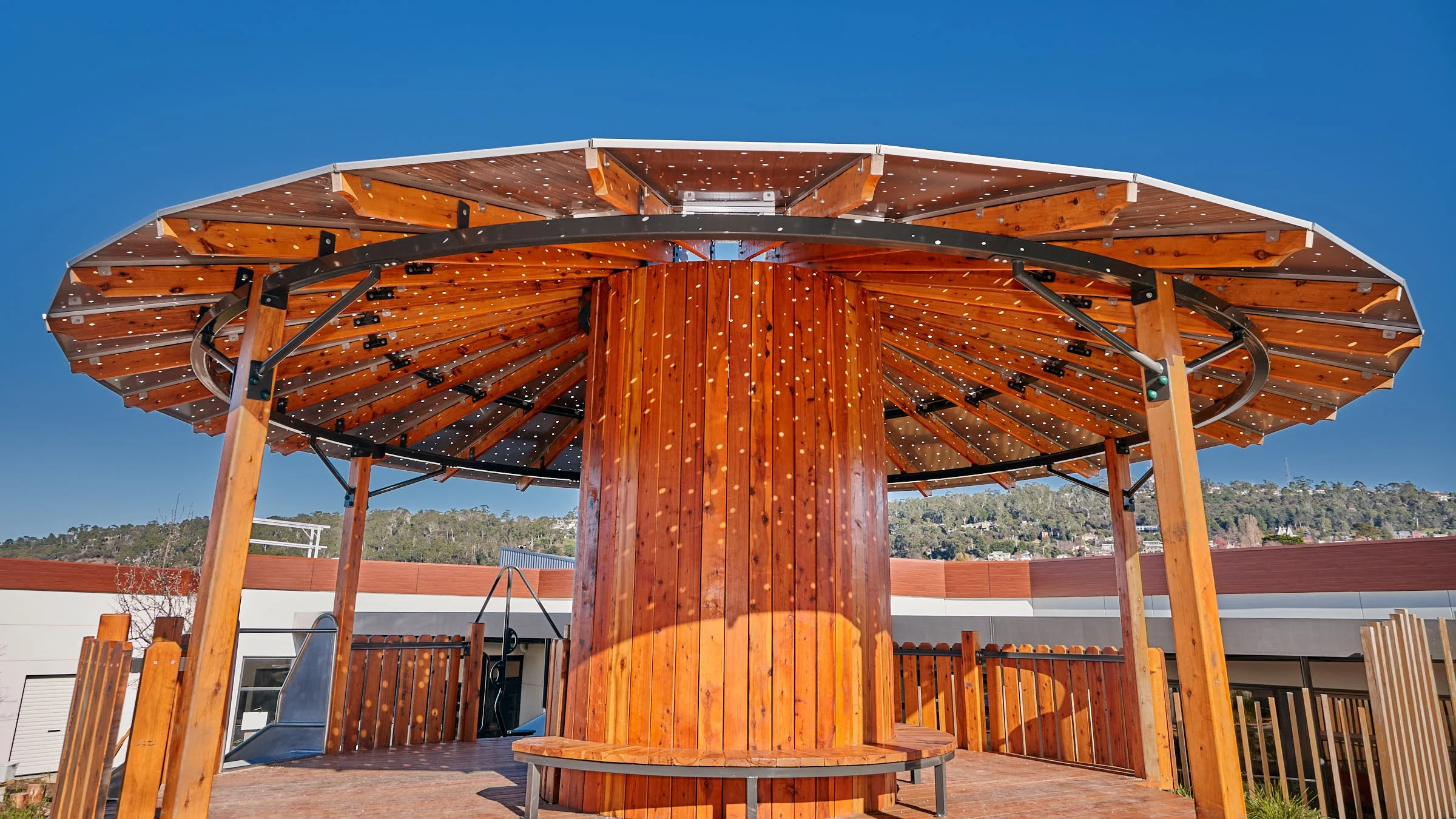 A large wooden outdoor structure with a circular base and a wide, sloped roof, situated on a rooftop with a clear blue sky and distant hills in the background.