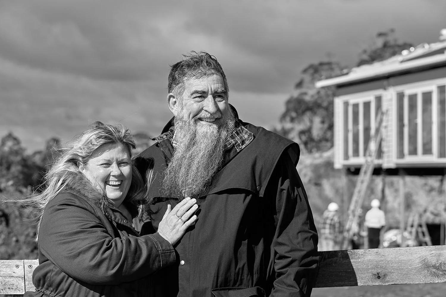 A smiling woman and a smiling man with a long beard, standing outdoors near a wooden fence, with a house and trees in the background, captured in black and white.