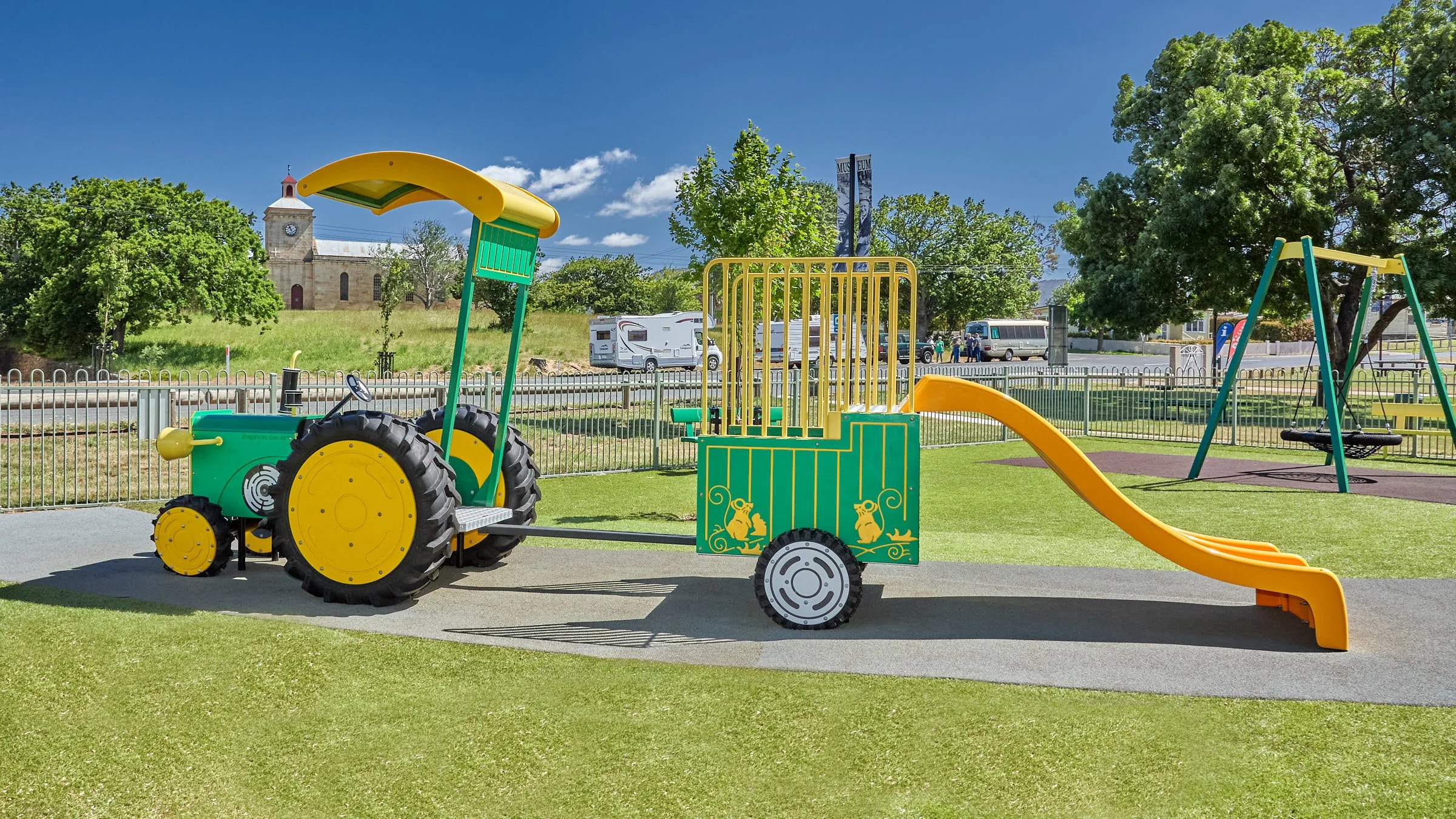 Colorful playground equipment shaped like a tractor with a slide in a park with green grass, trees, a fence, and a blue sky.