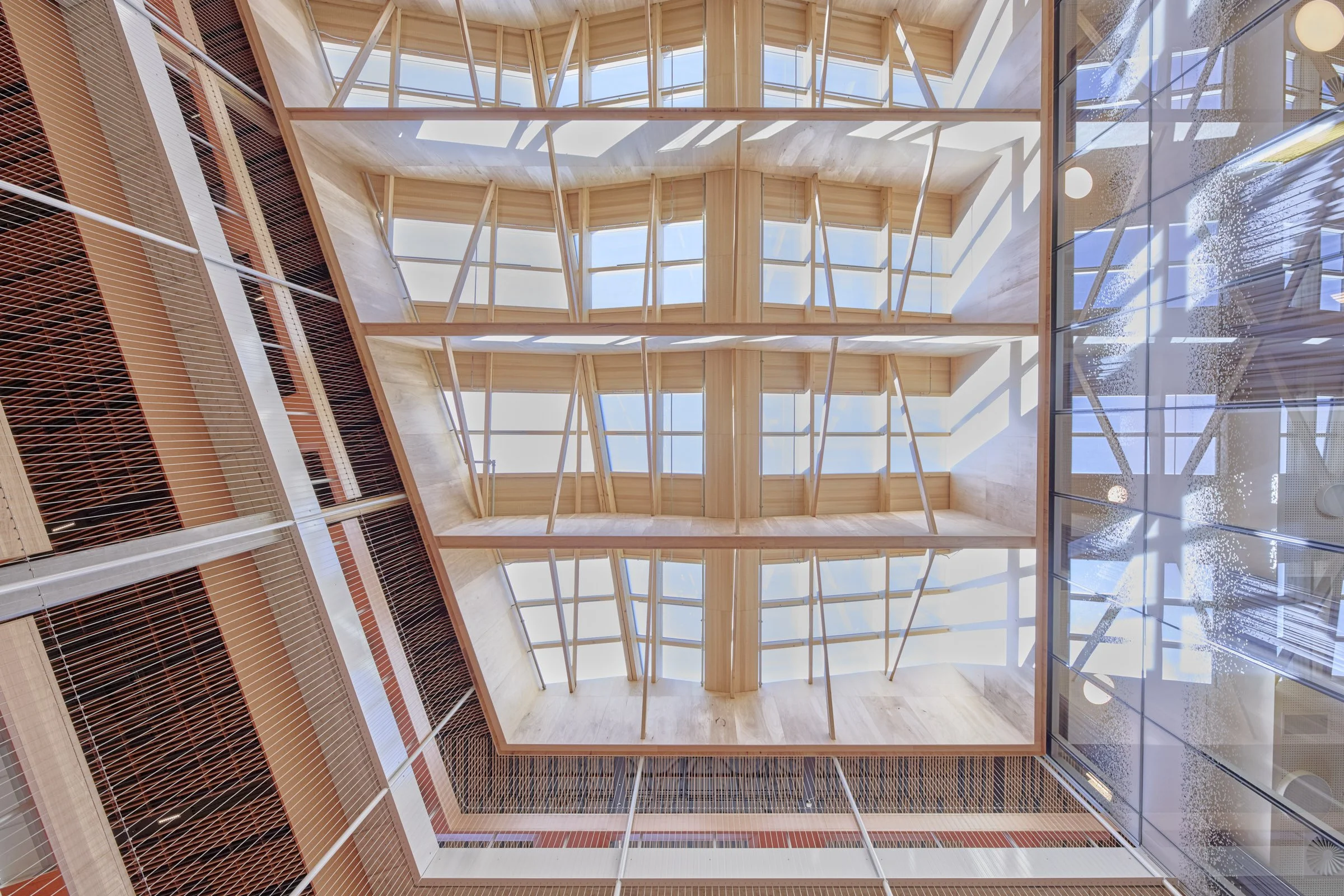 Looking up at the ceiling of a modern indoor atrium with large windows, wooden beams, and a glass roof allowing natural light.