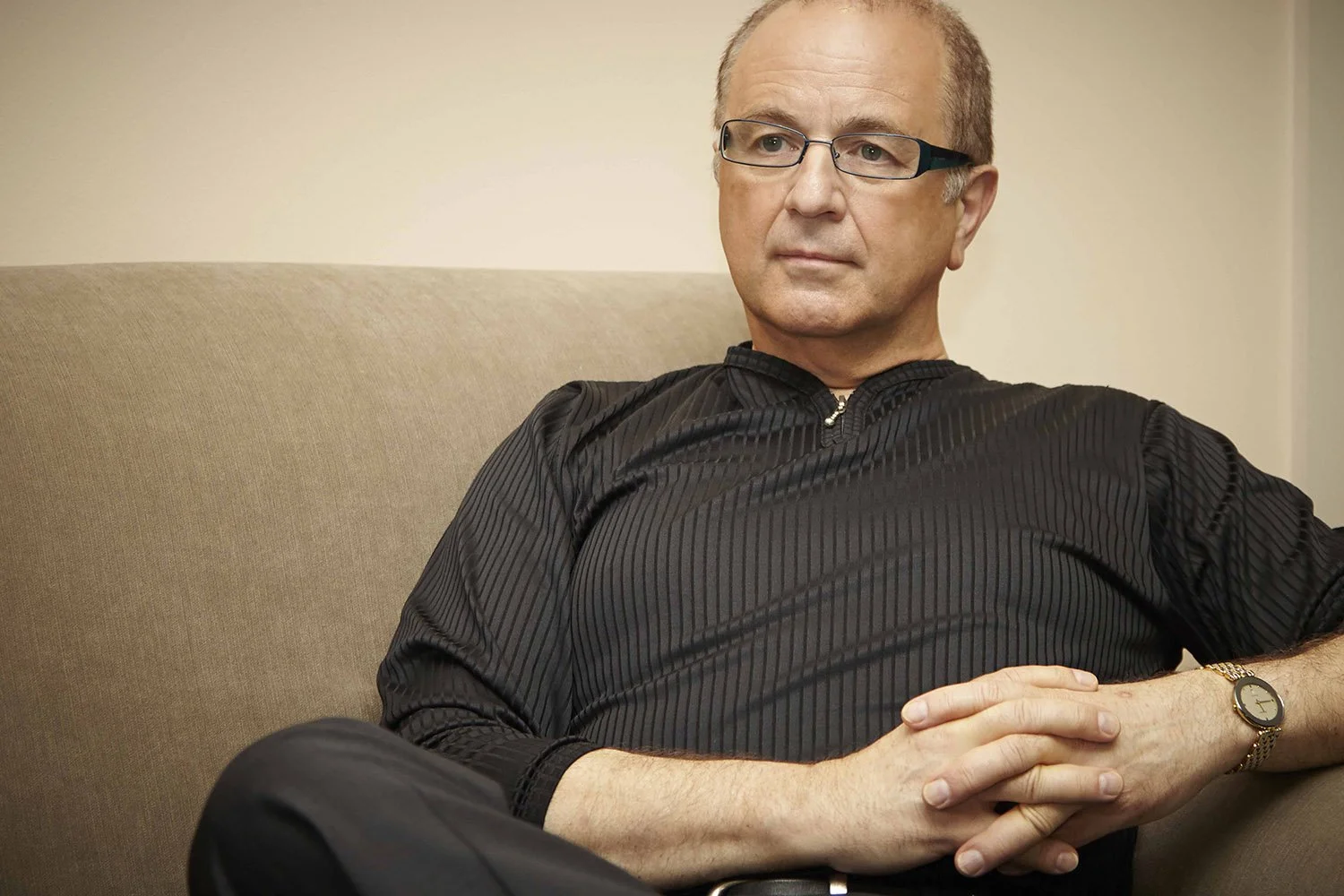 A middle-aged man with glasses, dressed in a black striped shirt, sitting on a beige sofa, looking contemplative with hands clasped over his stomach, wearing a wristwatch.