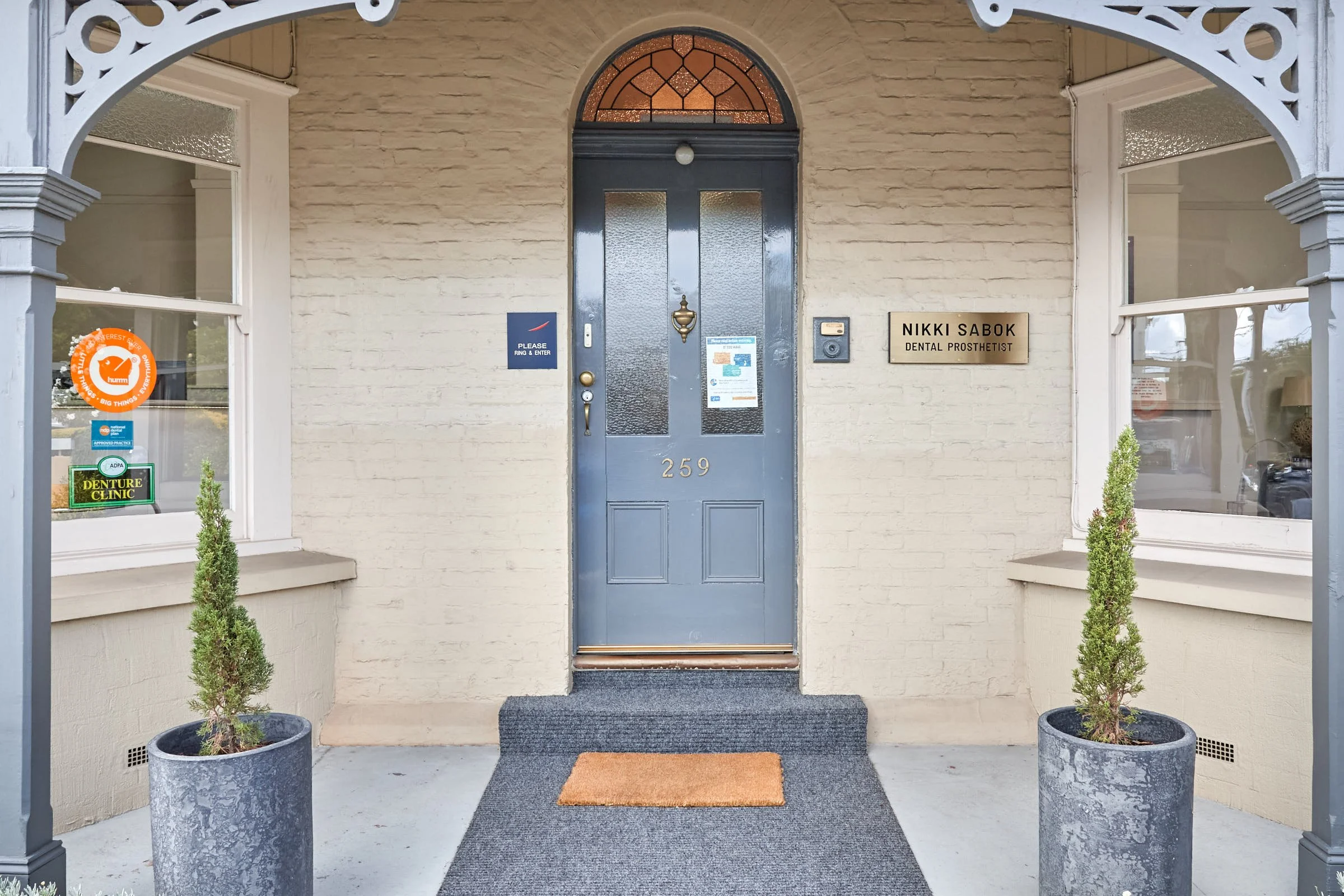The entrance to a dental prosthetist's office with a blue door, house number 259, flanked by two potted small trees. The door has glass panels and a potted plant plaque on the right side, with a sign that reads 'Nikki Sabok Dental Prosthetist'. There