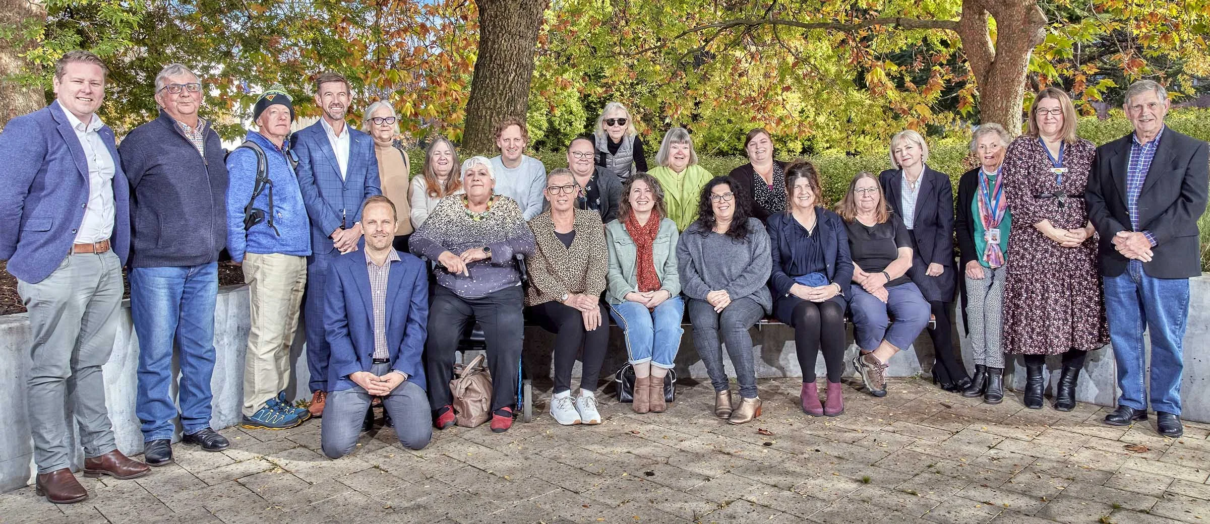 Group of people posing outdoors in front of trees with fall foliage, some sitting and others standing.